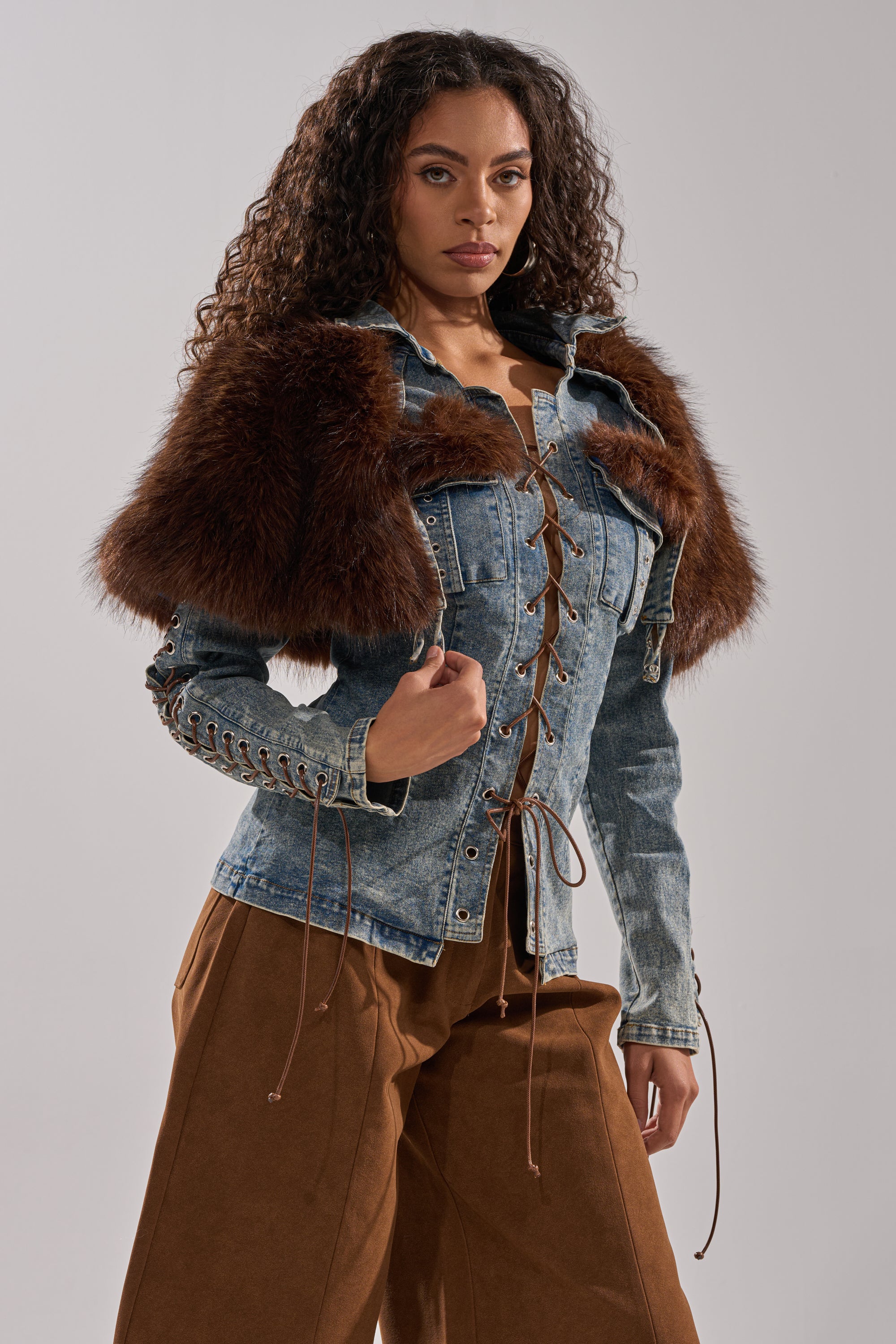 A woman with curly hair wears the TRY ME BABY LACE UP DENIM TOP, featuring corset-inspired lace-up front and faux fur panel shoulders, paired with brown wide-leg pants, posing confidently against a plain gray background.