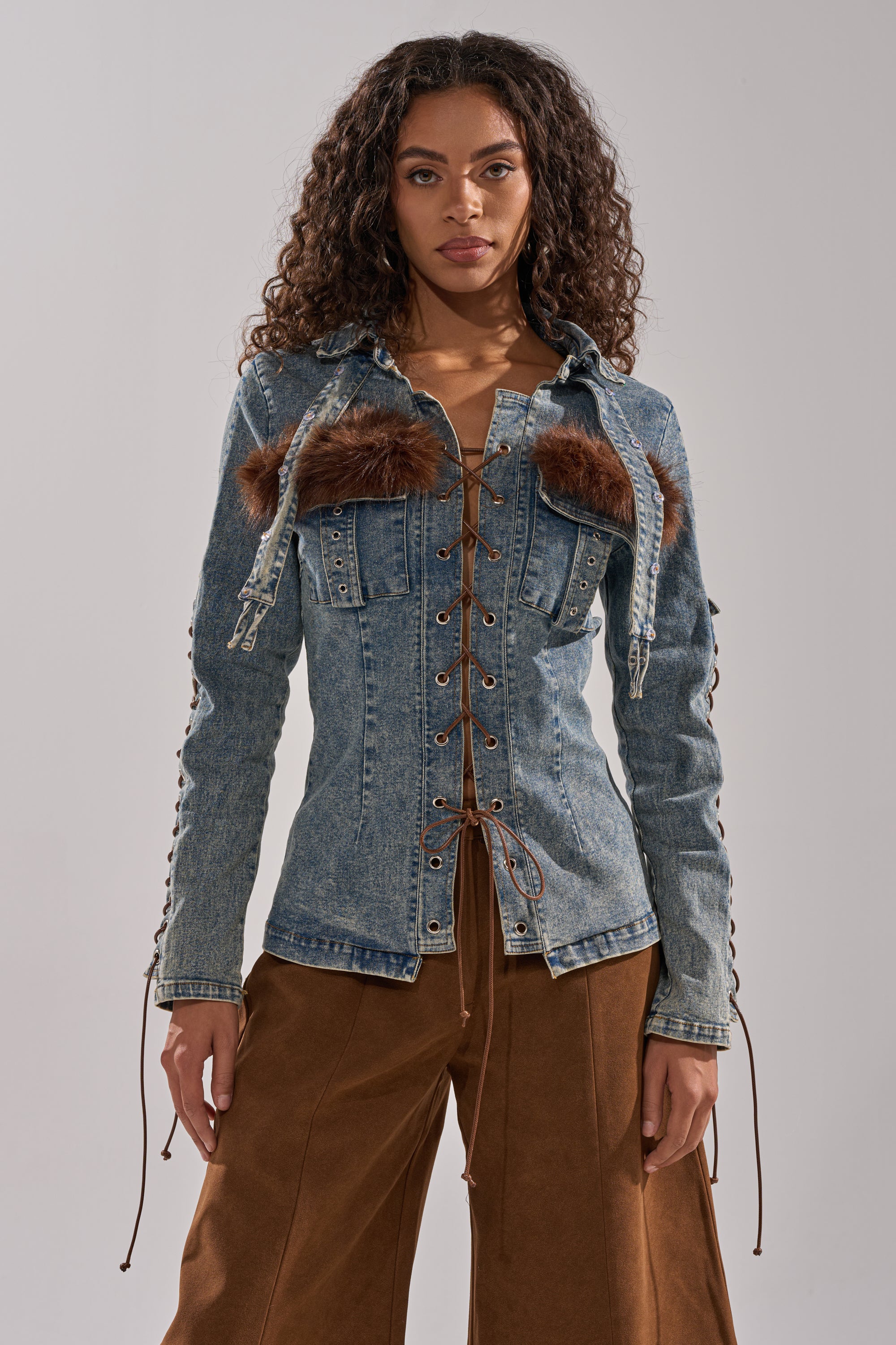A woman with curly hair wears the TRY ME BABY LACE UP DENIM TOP, featuring lace-up details and faux fur panels, paired with brown wide-leg pants, standing against a neutral background.