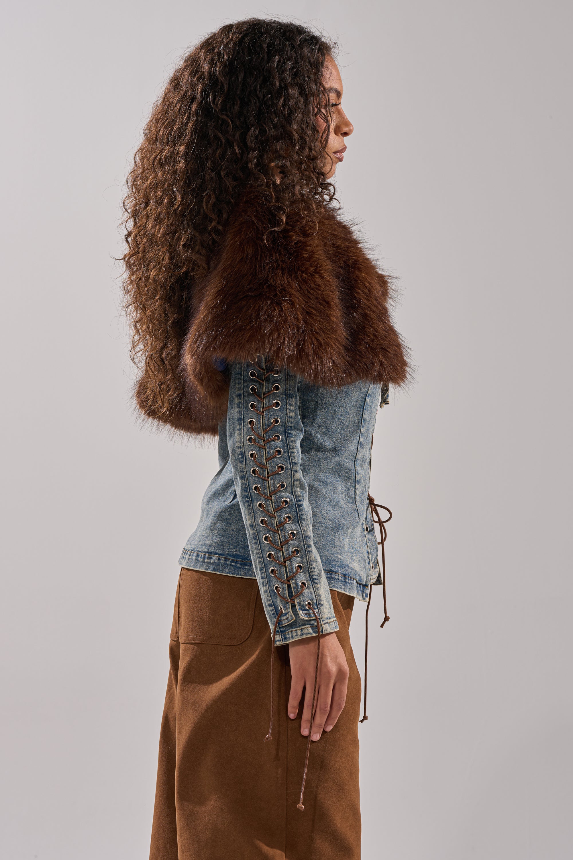 A woman with curly hair stands in profile against a plain background, wearing the TRY ME BABY LACE UP DENIM TOP with lace-up sleeves and a faux fur panel collar, paired with a brown skirt.