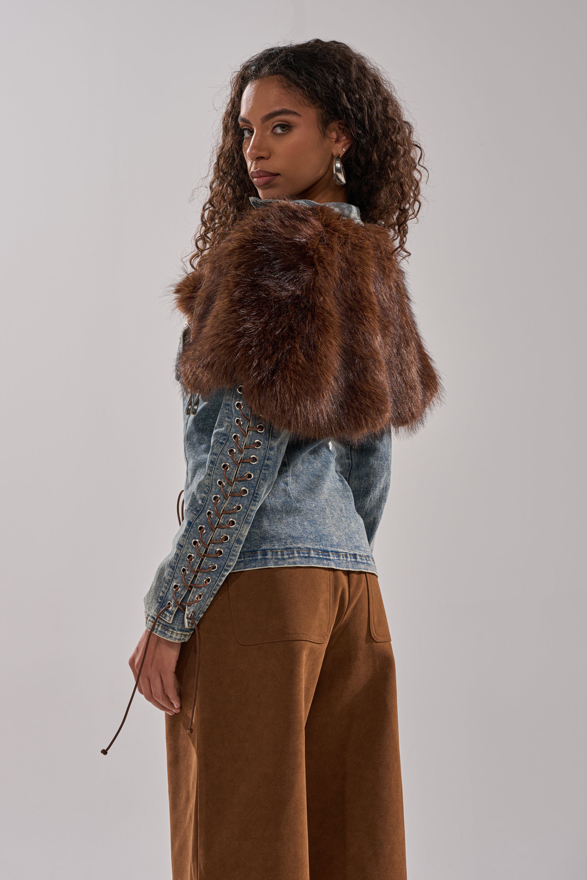 A woman with curly hair looks over her shoulder, wearing the TRY ME BABY LACE UP DENIM TOP featuring corset-inspired lace-up details and faux fur accents, paired with brown wide-leg pants against a plain background.