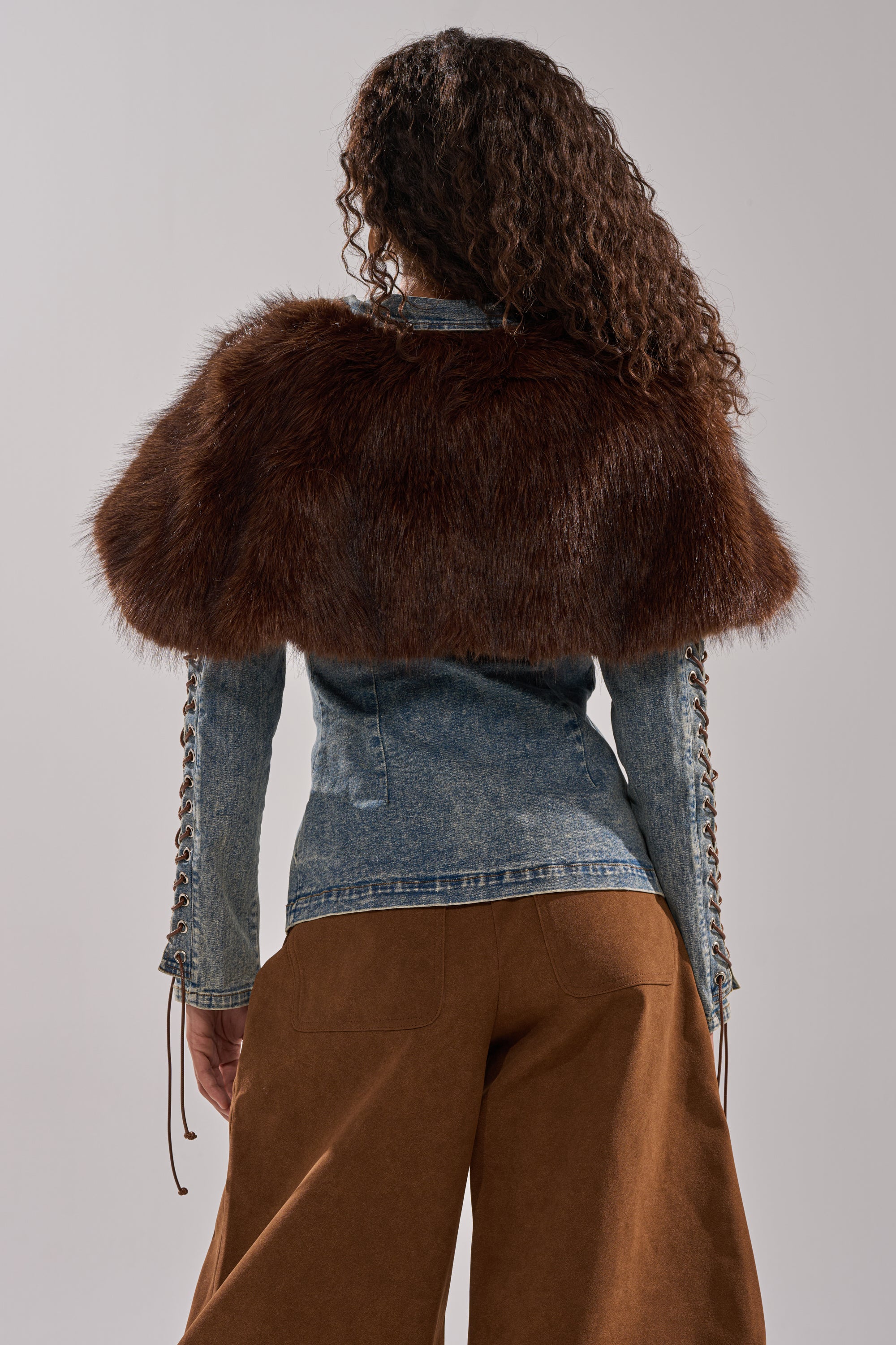 A person with long curly hair is shown from the back, wearing the TRY ME BABY LACE UP DENIM TOP with lace-up sleeves, a faux fur panel over their shoulders, and wide-legged brown pants against a plain neutral background.