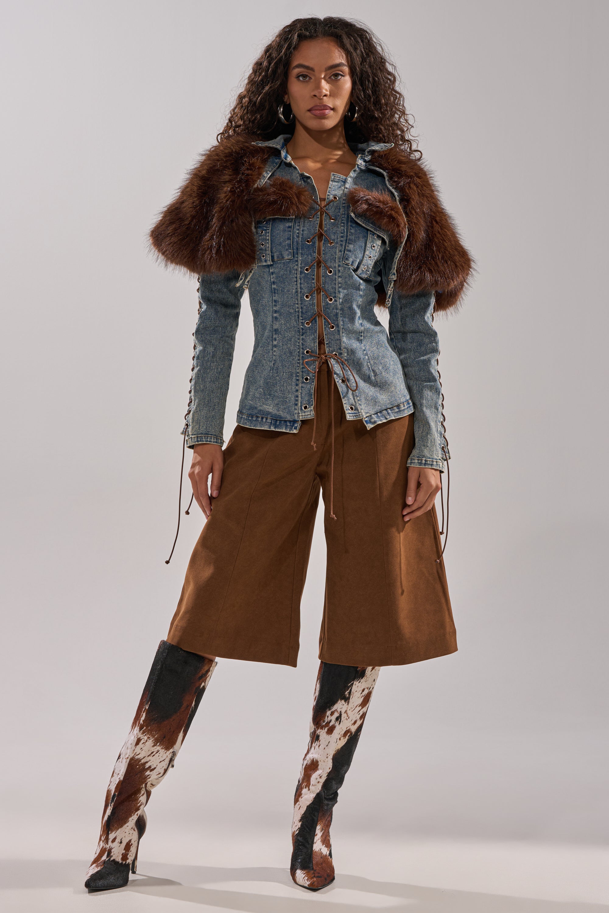 A woman faces forward in the TRY ME BABY LACE UP DENIM TOP with brown faux fur panels, paired with brown culotte pants and cowhide knee-high boots, set against a plain light gray background.