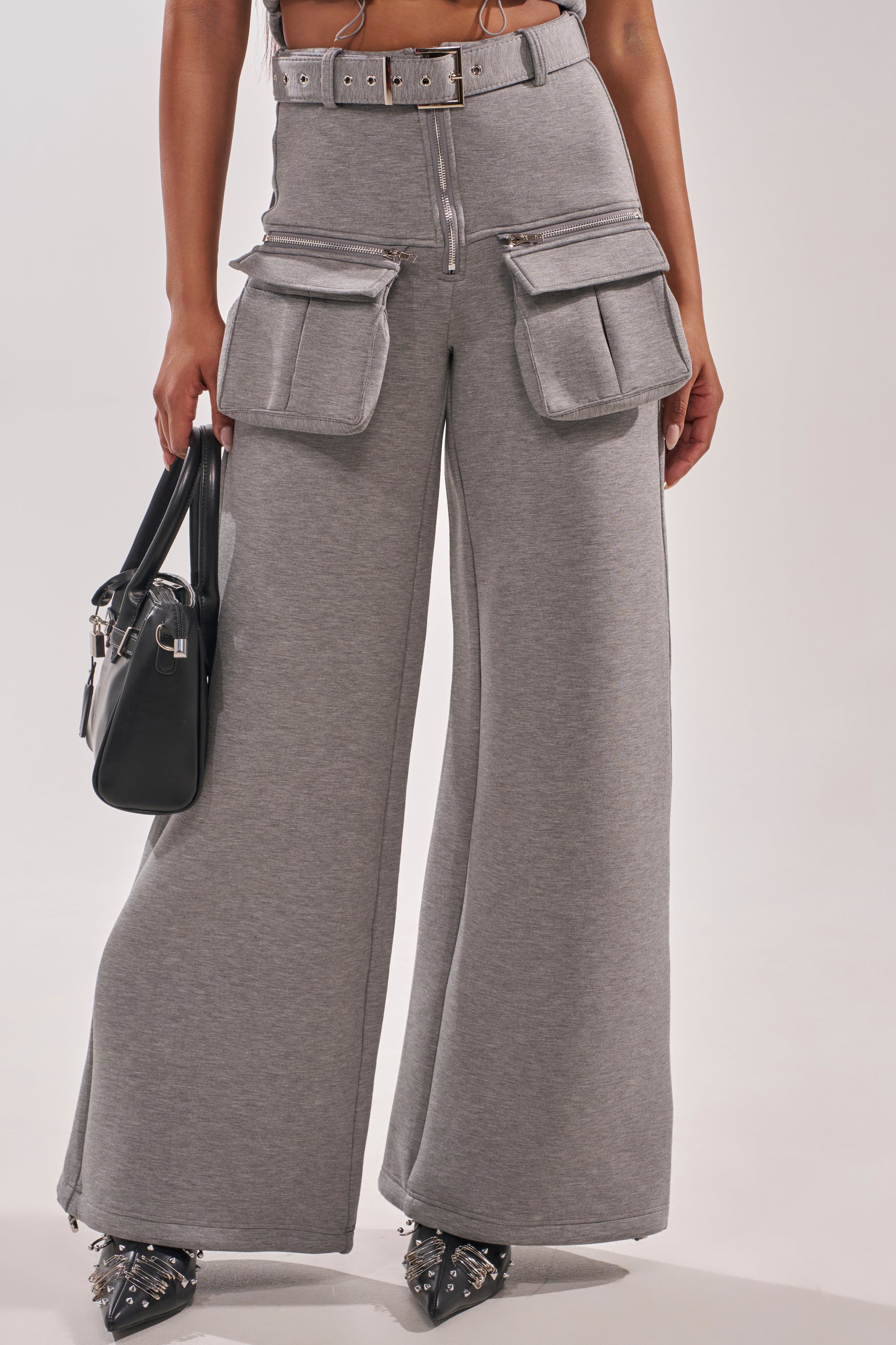 A person wearing the THAT GIRL SCUBA CARGO PANT, featuring high-waisted wide-leg grey design, large front flap pockets, and a matching belt, holds a black handbag. Only the lower body is visible. 1