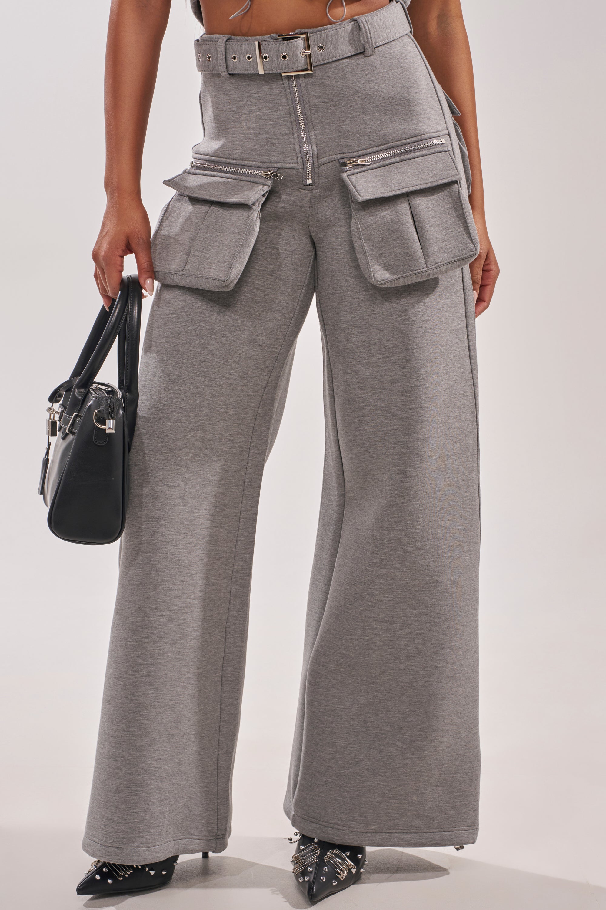 Wearing THAT GIRL SCUBA CARGO PANT—high-waisted grey cargo pants with big pockets, zippers, and a belt—styled with black pointed studded shoes and a black handbag for a chic, functional look. 2