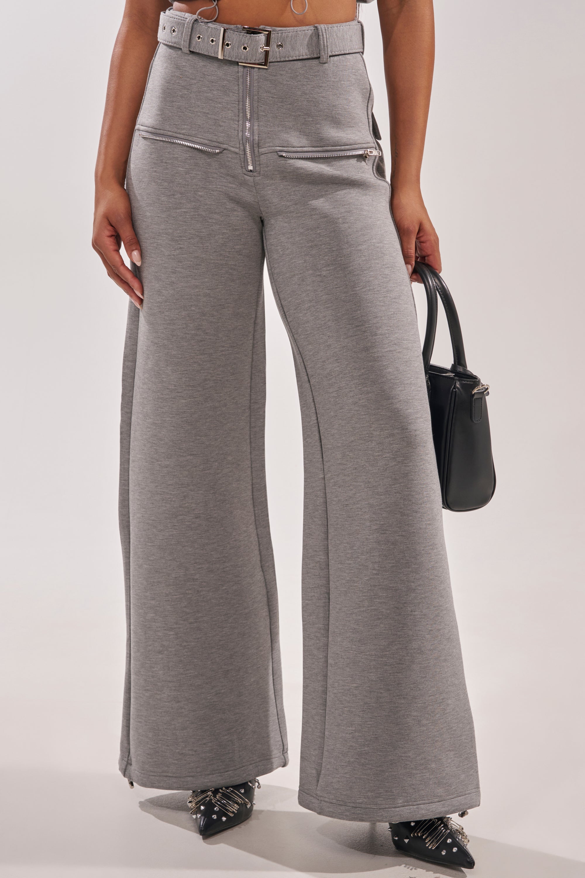 Wearing the THAT GIRL SCUBA CARGO PANT—high-waisted, wide-leg grey pants with a matching belt and zippered pockets—the person holds a small black handbag. Only the lower half of their body is shown. 3