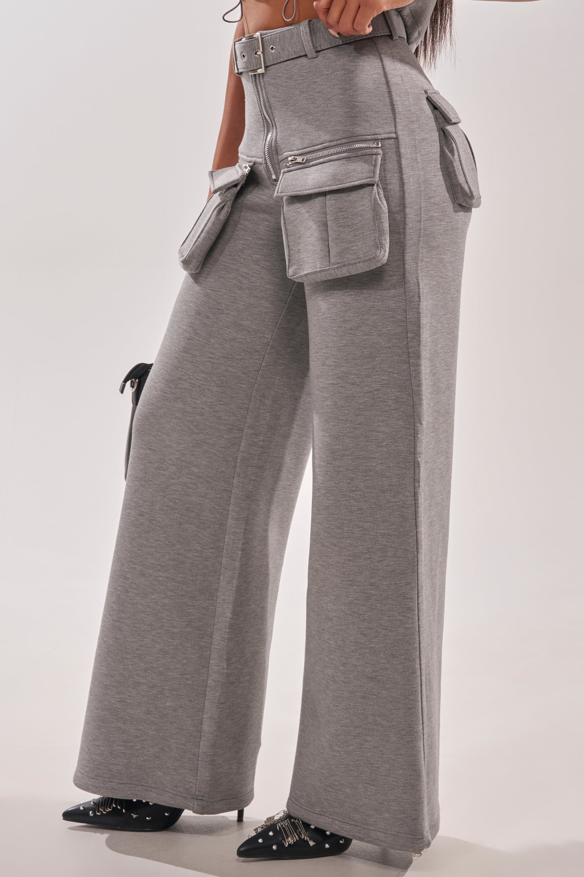 Wearing the THAT GIRL SCUBA CARGO PANT in high-waisted grey with multiple large pockets and a belt, paired with pointed black heels with silver studs. Their hands are in or near the cargo pockets. 4
