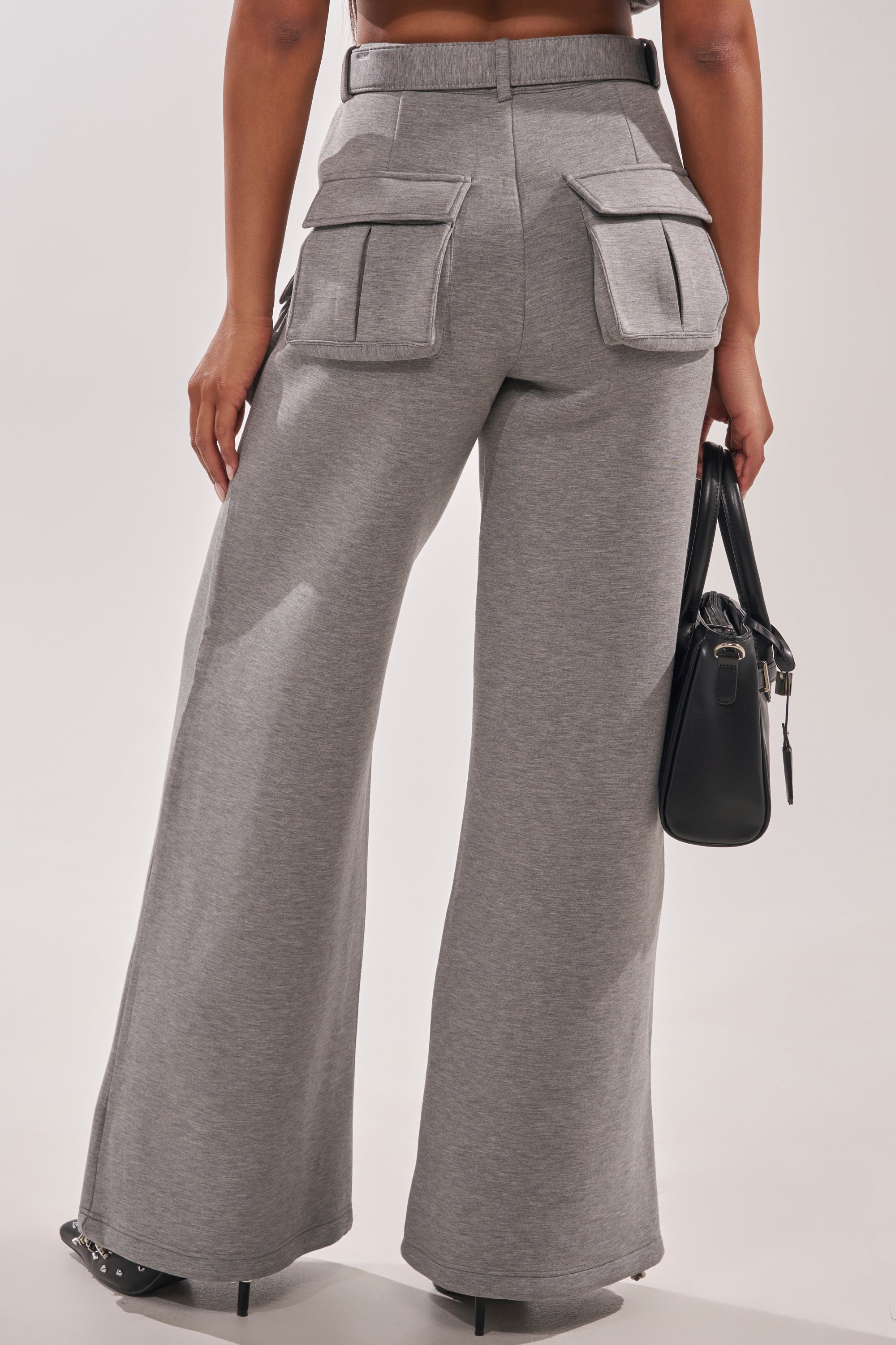 A person models the THAT GIRL SCUBA CARGO PANT in gray—high-waisted, wide-leg style with large back and side pockets. The focus is on the waist-down, back view, and they're holding a black handbag. 5