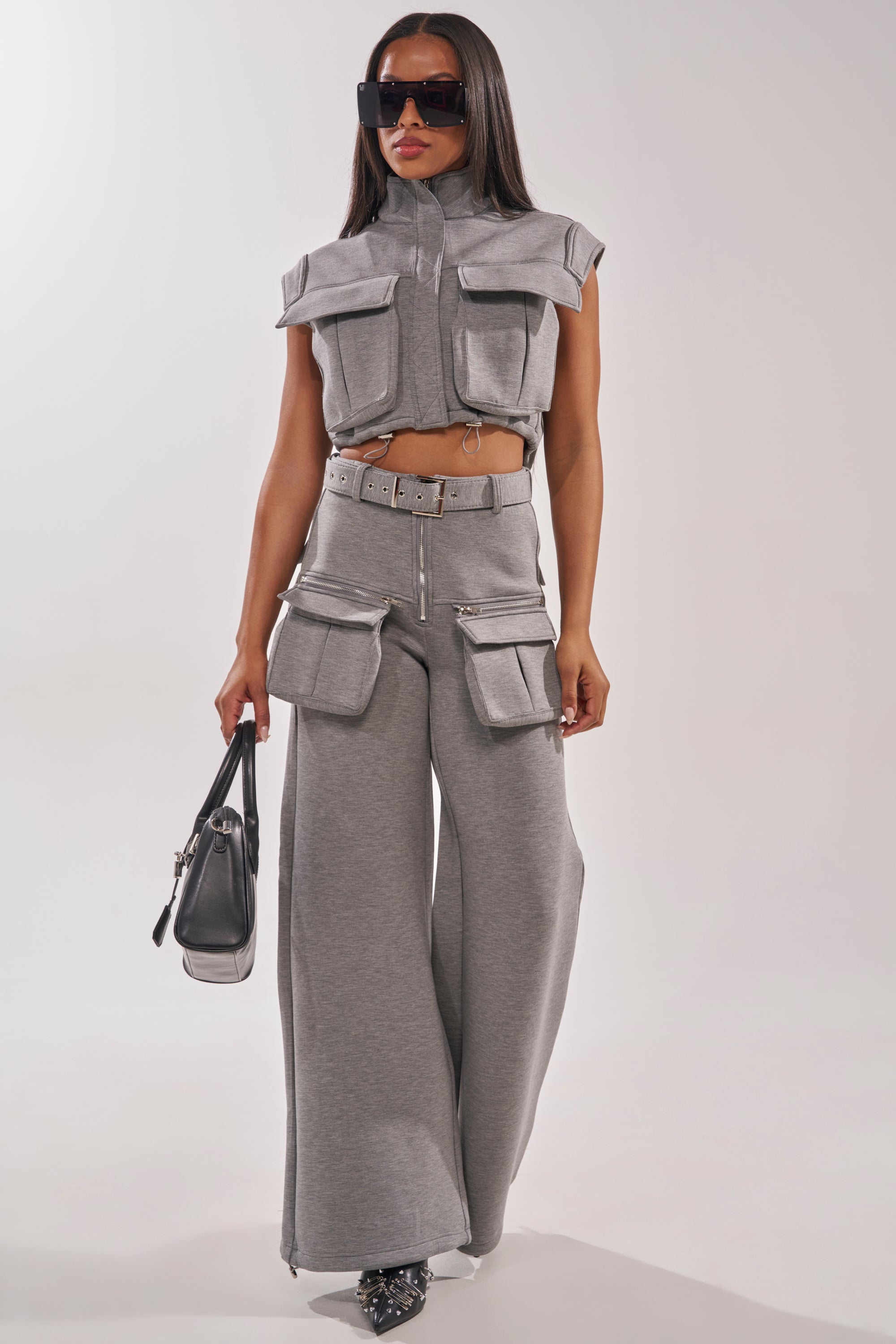 A woman poses in THAT GIRL SCUBA CARGO PANT featuring large pockets, paired with a sleeveless cropped utility jacket, oversized sunglasses, studded pointed shoes, and a black handbag against a plain background. 6