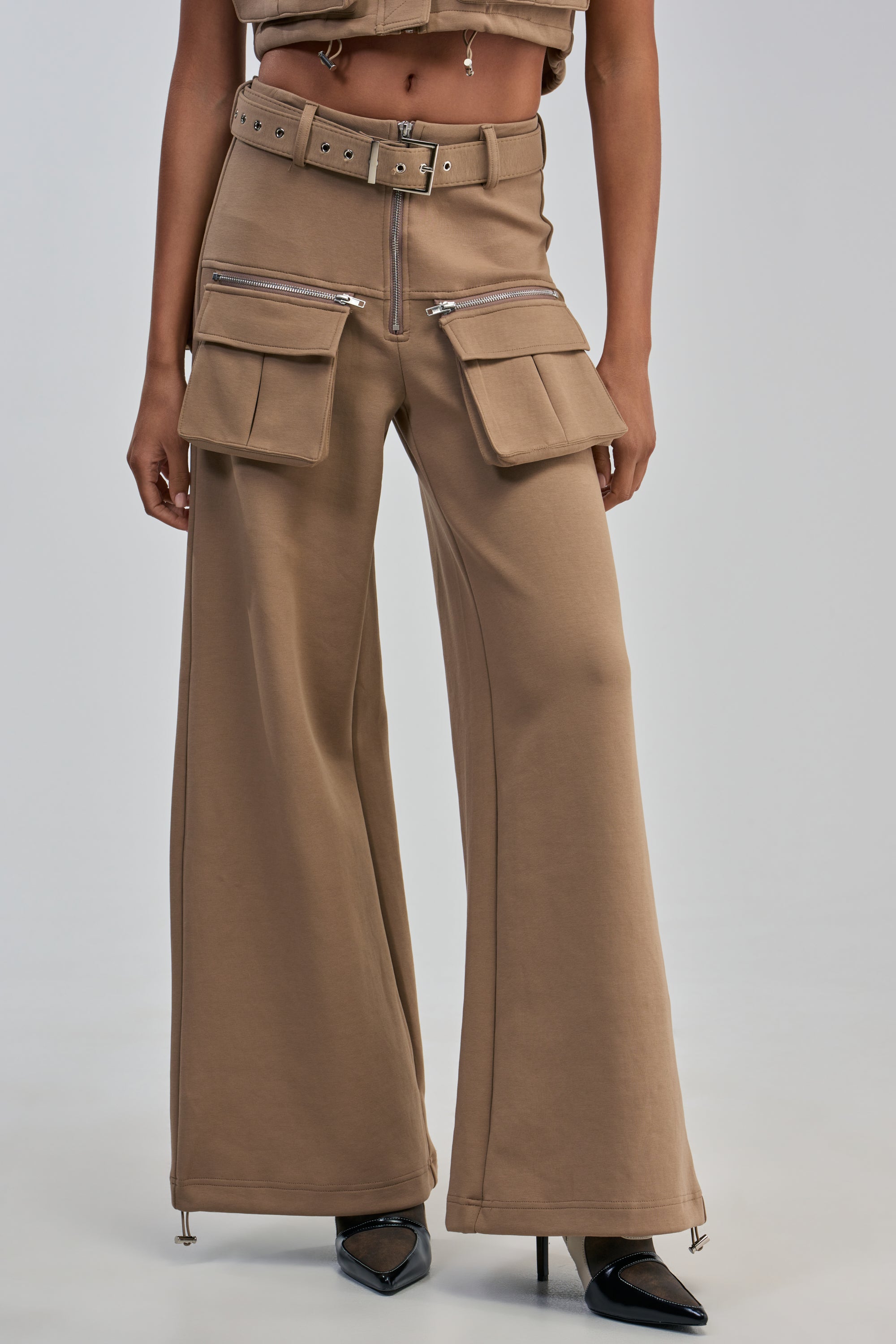 Wearing the THAT GIRL SCUBA CARGO PANT IN TAUPE—high-waisted with a wide-leg silhouette, large cargo pockets, zippers, and a matching belt—paired with pointed black heels, this look channels utility-chic style with a modern edge.