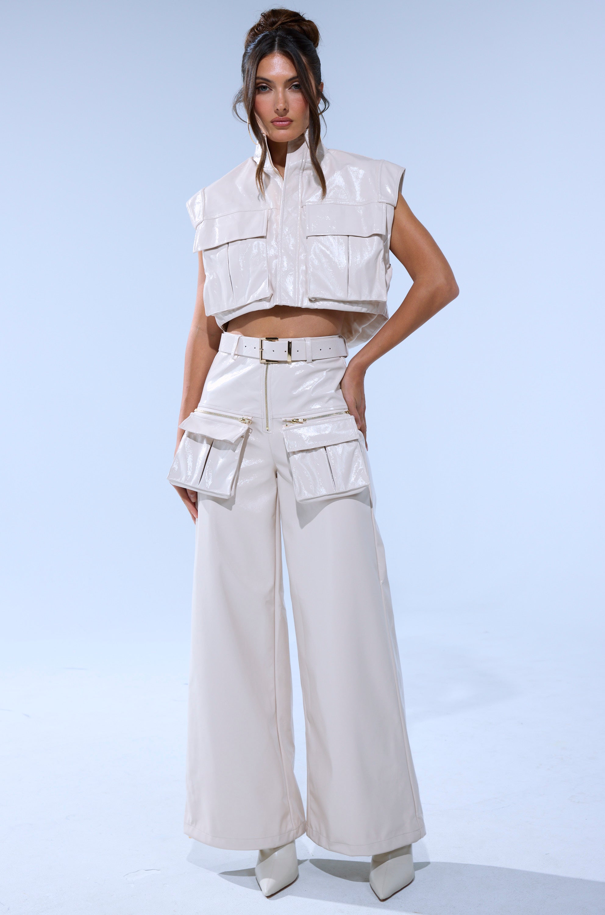 A woman poses in SHOW STOPPER FAUX LEATHER CARGO TROUSER, styled in shiny light beige with a sleeveless crop vest, wide-leg silhouette, high-rise fit, and large front pockets. She completes the look with white pointed-toe shoes.
