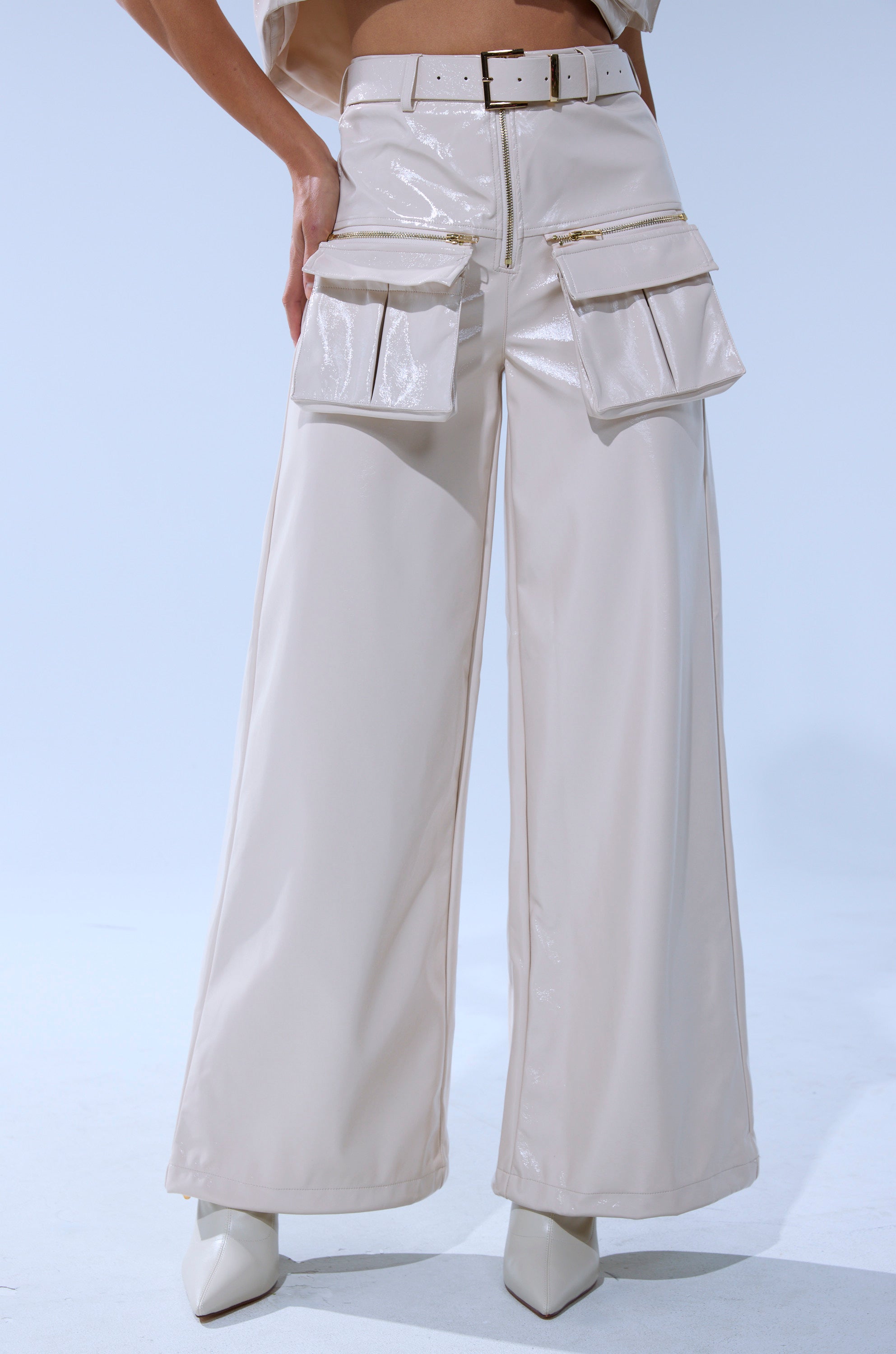 A person models the SHOW STOPPER FAUX LEATHER CARGO TROUSER—cream, shiny, high-rise with wide legs and large front pockets—styled with a matching belt and pointed-toe shoes against a plain light background.