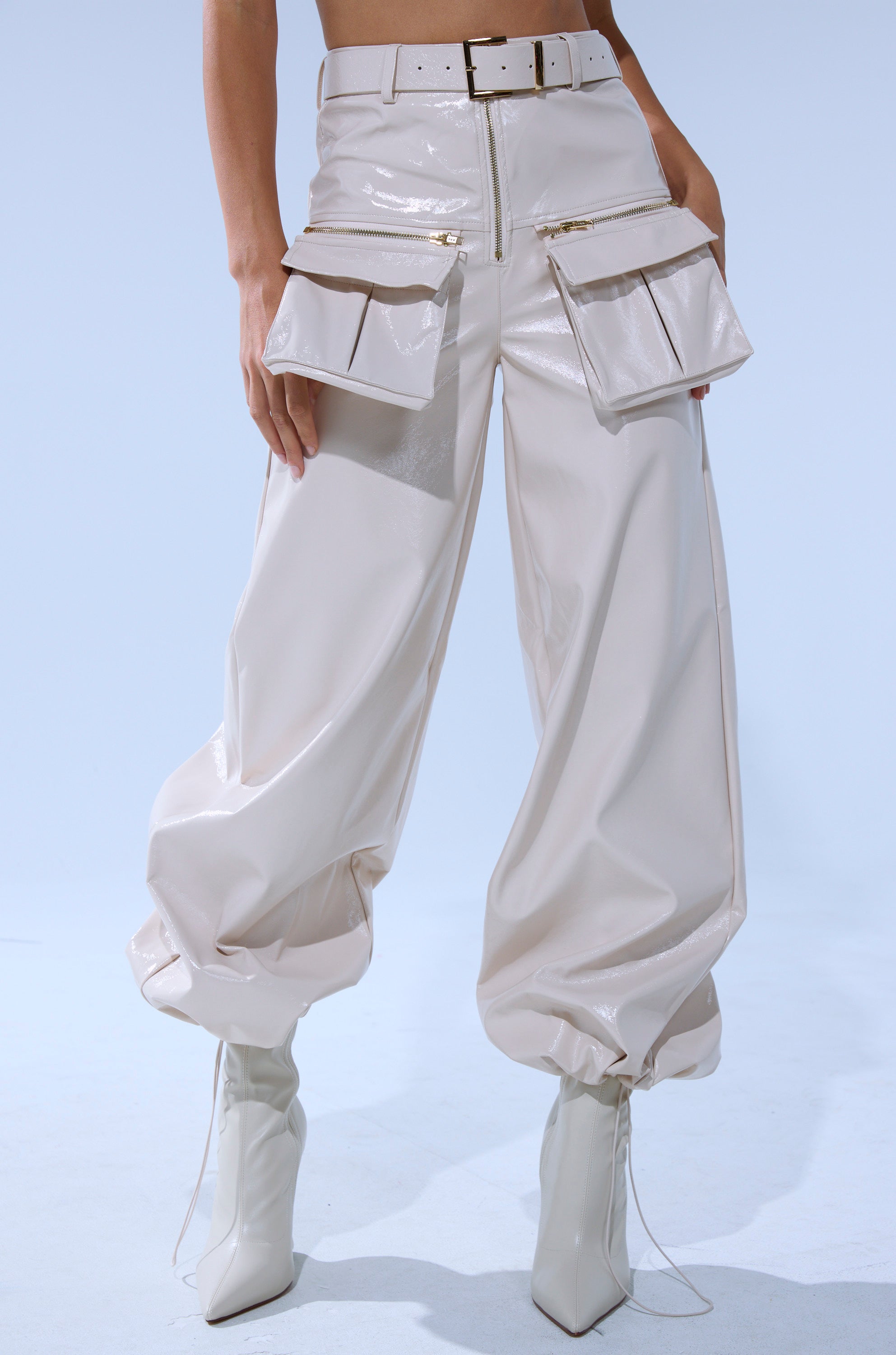 A person wears the SHOW STOPPER FAUX LEATHER CARGO TROUSER: shiny, cream, high-rise cargo pants with large zippered pockets, a matching belt, gathered ankle cuffs, wide-leg fit, and pointed-toe heeled boots.