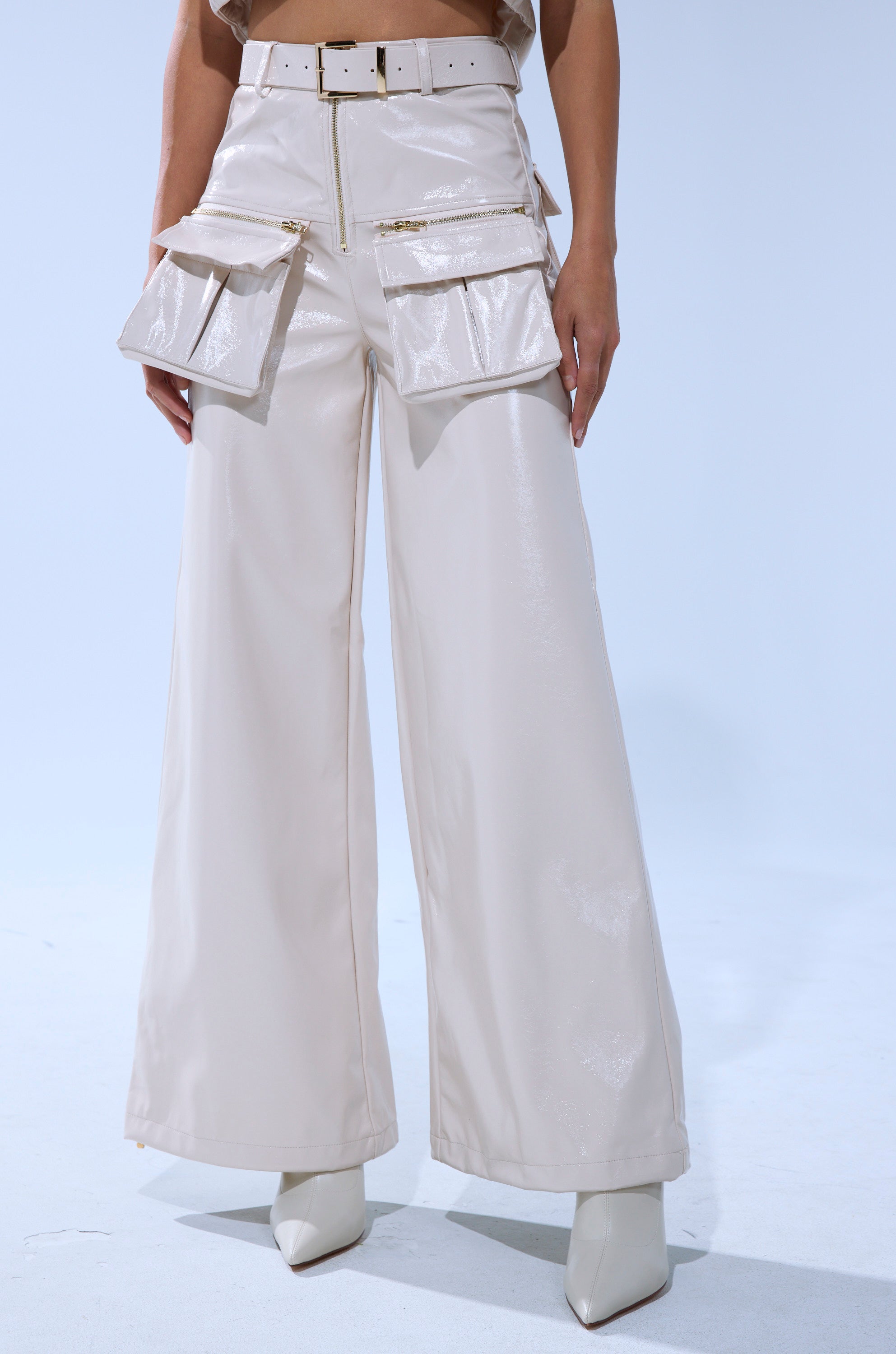 A person wears the SHOW STOPPER FAUX LEATHER CARGO TROUSER—shiny, cream, high-rise wide-leg pants with large front pockets—paired with matching heels against a light blue background.