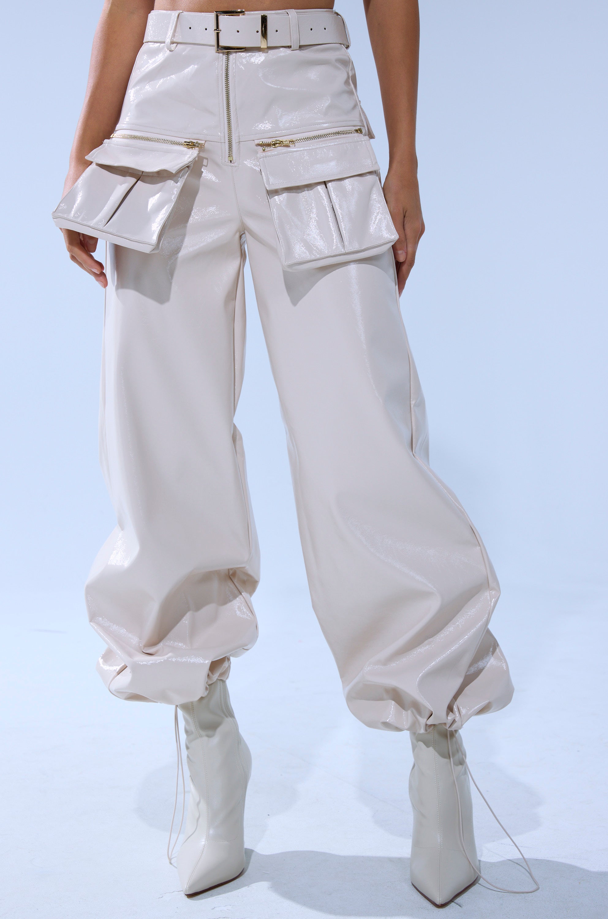 Someone models the SHOW STOPPER FAUX LEATHER CARGO TROUSER—shiny, cream, wide-leg with large pockets and a matching belt—styled with white pointed-toe heeled boots against a plain light blue background.