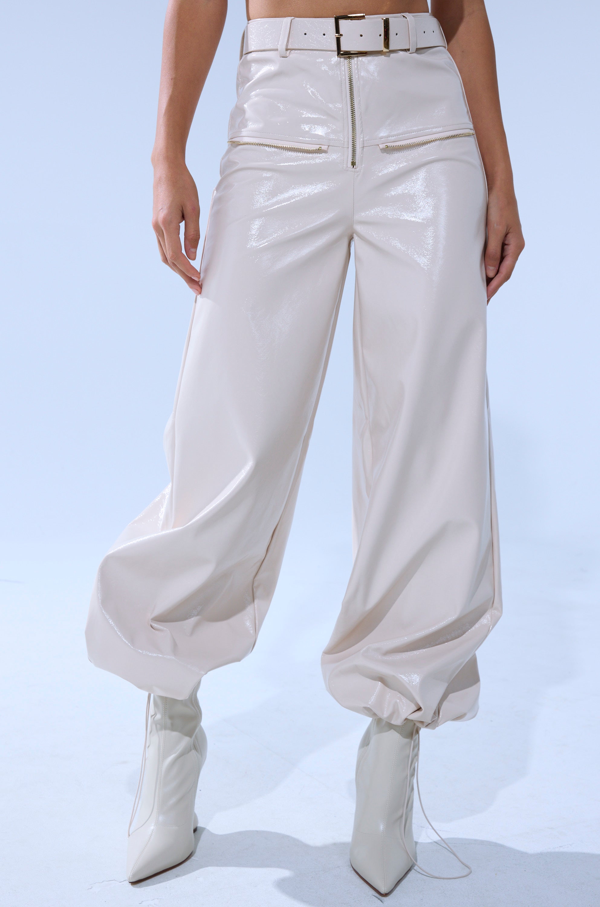 A person wears the SHOW STOPPER FAUX LEATHER CARGO TROUSER—high-waisted, shiny beige, with a matching belt and gathered ankles—paired with white pointed-toe boots, standing against a light blue background.