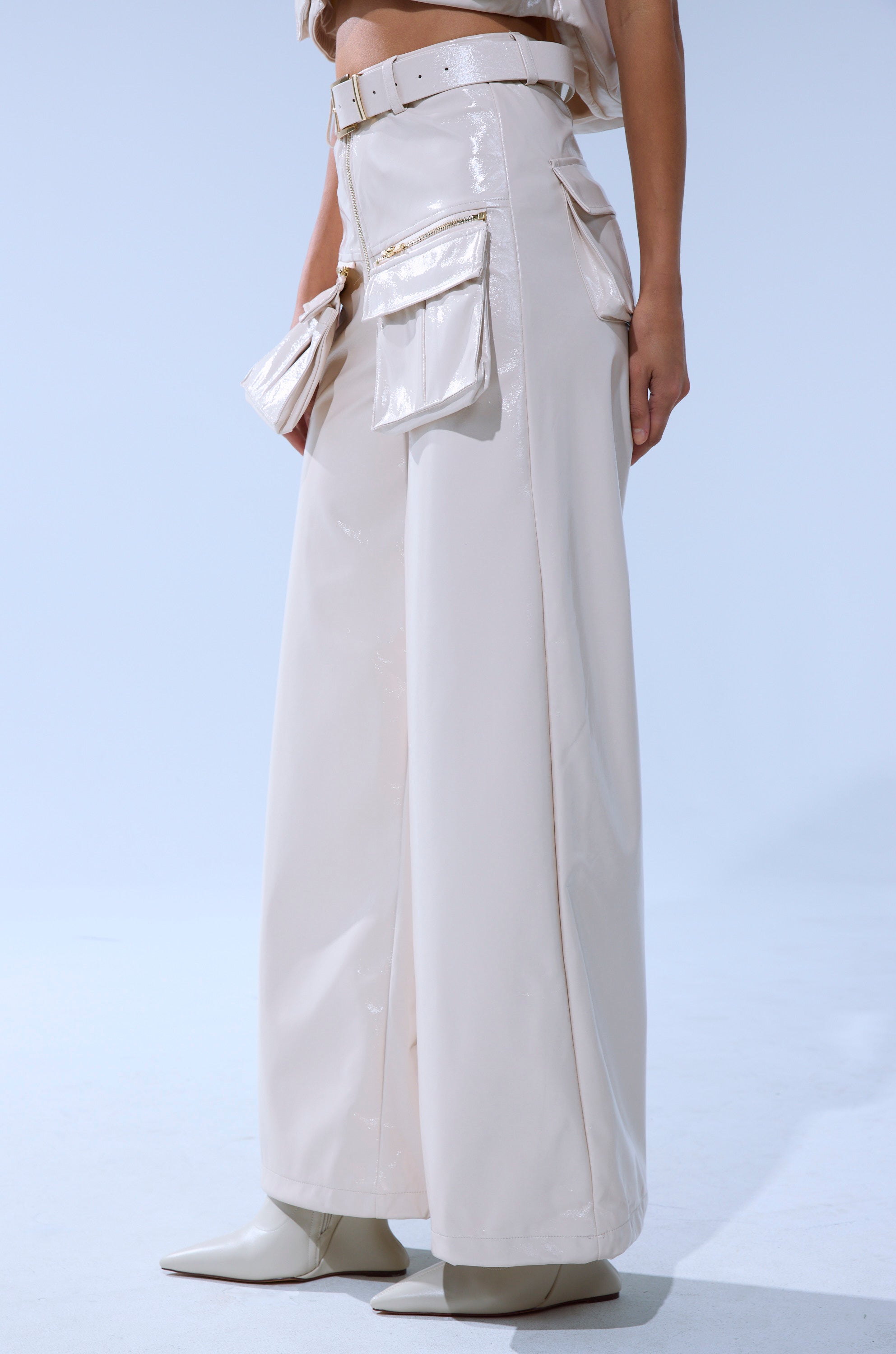 A person wears the SHOW STOPPER FAUX LEATHER CARGO TROUSER—glossy cream, high-rise, wide-leg style with large side pockets and a matching belt—paired with pointed-toe boots for a modern look against a plain background.