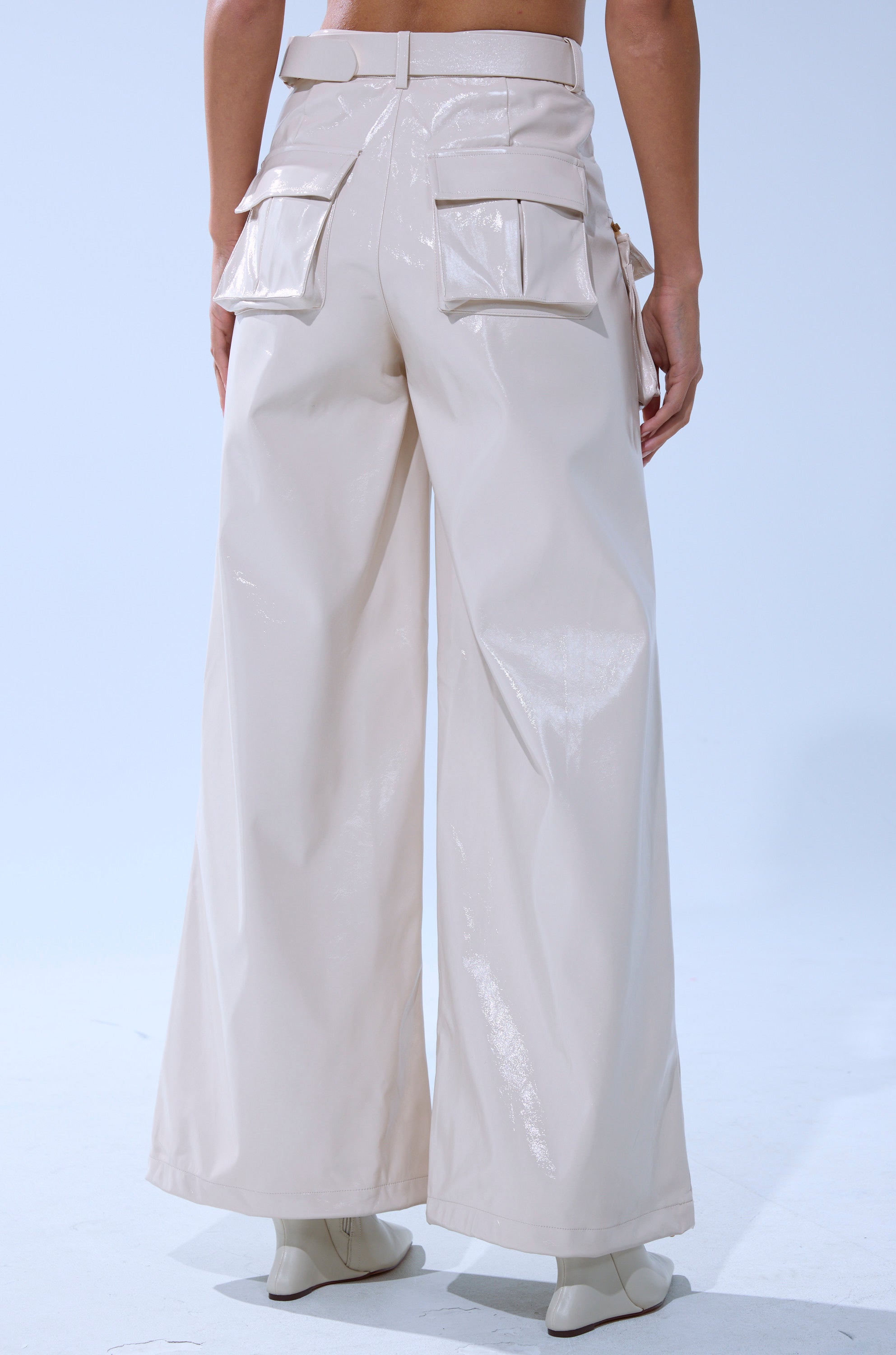 A person wears SHOW STOPPER FAUX LEATHER CARGO TROUSER in a wide-leg fit with glossy finish and large back flap pockets, paired with cream heeled boots. Background is plain and light-colored.