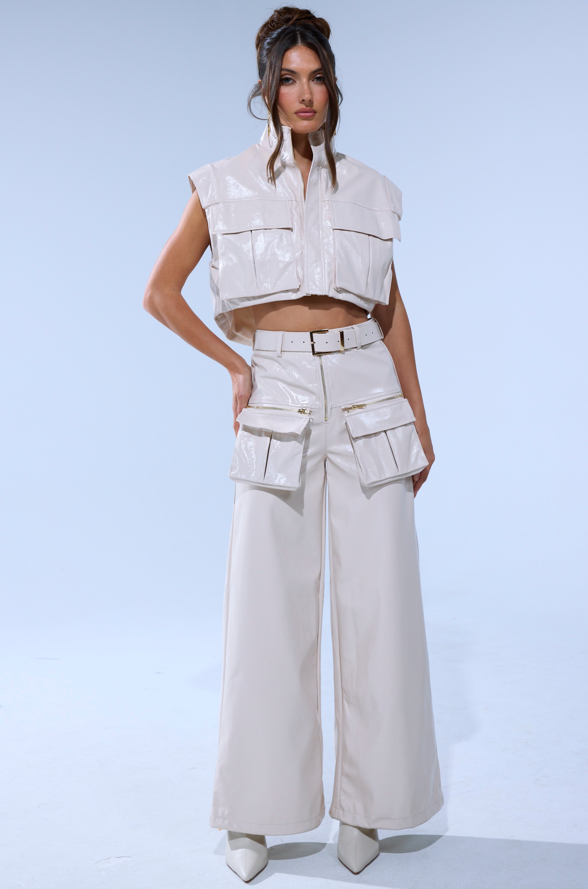 A woman poses against a light background, wearing SHOW STOPPER FAUX LEATHER CARGO TROUSER—a shiny, sleeveless light beige cropped jacket with front pockets and matching high-rise, wide-leg cargo trousers with oversized pockets and pointed-toe shoes.
