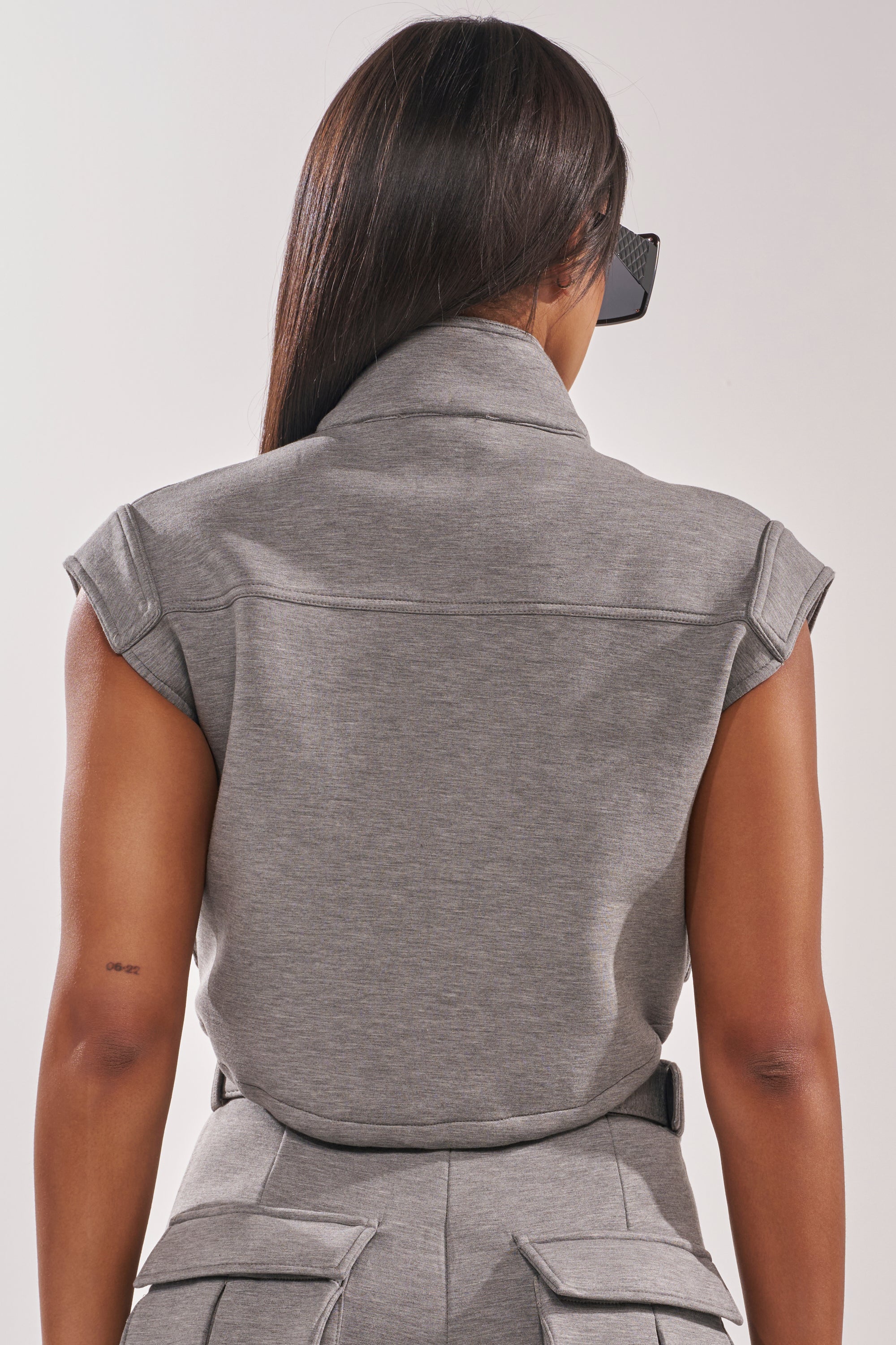 A woman with long straight hair and large sunglasses is shown from the back, wearing the THAT GIRL SCUBA TOP in grey, styled as a short-sleeve collared jumpsuit with structured shoulders and cargo pockets. 3