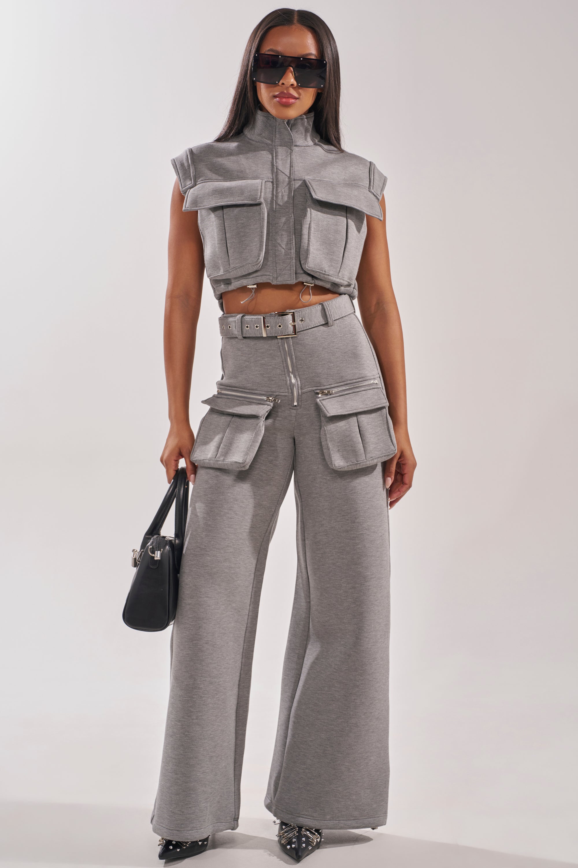 A woman wears the THAT GIRL SCUBA TOP, a grey knit sleeveless cargo blouse, paired with matching high-waisted wide-leg pants featuring large pockets. She styles it with long hair, sunglasses, a black handbag, and black pointed shoes. 4
