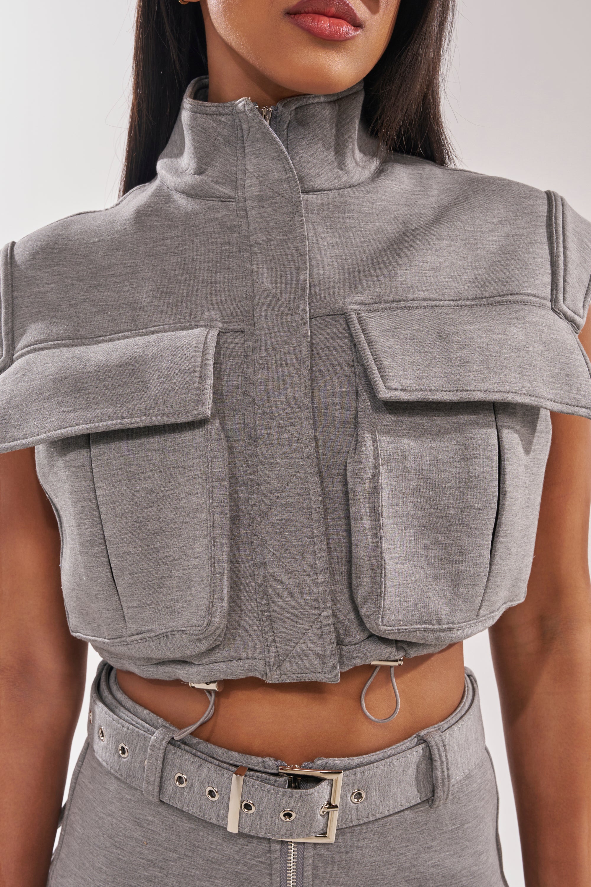 A woman wears the THAT GIRL SCUBA TOP—a grey mockneck sleeveless blouse styled as a cropped vest with large front pockets and a matching high-waisted belt. The soft, textured fabric gives this festival look a modern edge. Her face is partially visible. 6