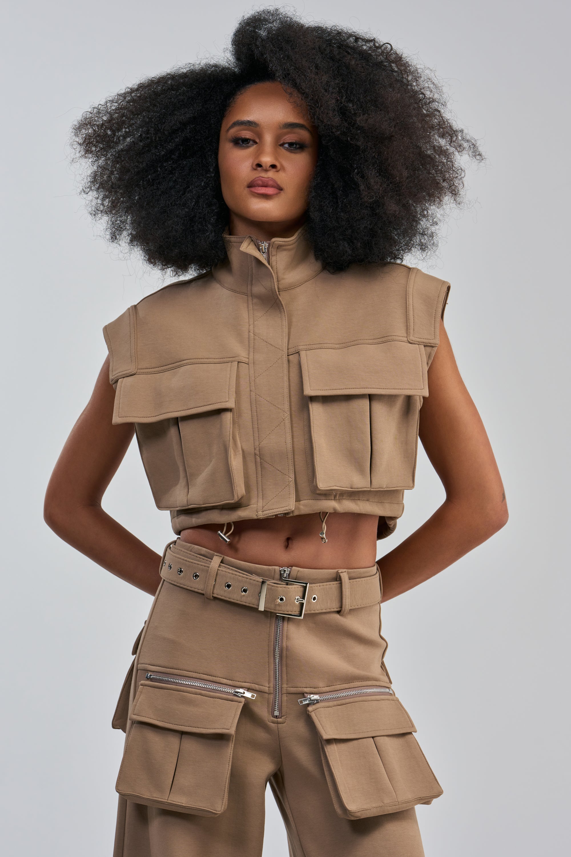 Wearing the THAT GIRL SCUBA TOP IN TAUPE styled as a sleeveless crop jacket with matching utility pants featuring large pockets and silver zippers, a woman with natural afro hair poses confidently against a plain light background, embodying effortlessly cool streetwear-inspired casual style.