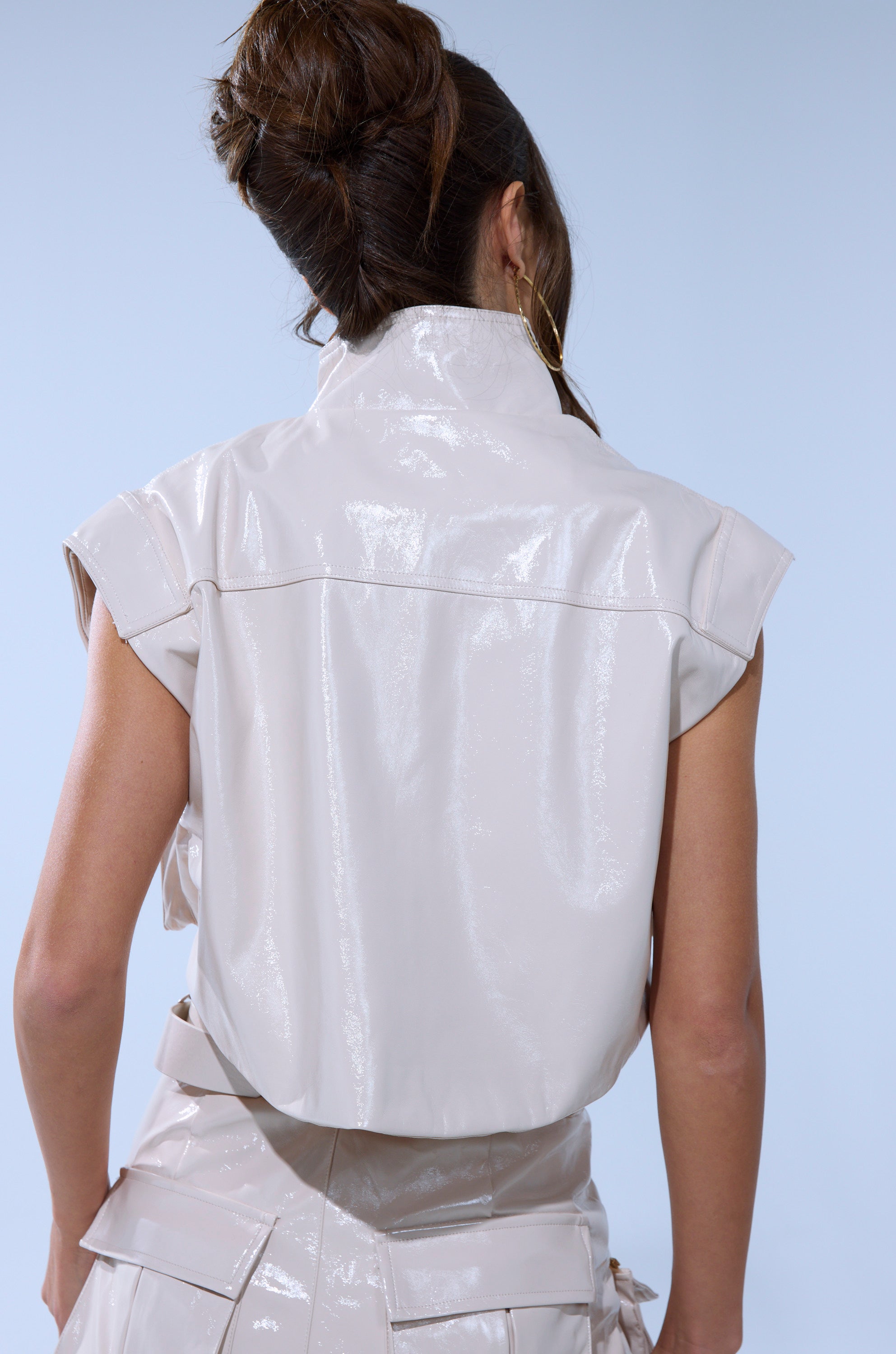 A woman with brown hair in an updo is seen from behind, wearing the SHOW STOPPER FAUX LEATHER ZIP UP TOP and a matching belted skirt in shiny light beige, set against a light blue background.