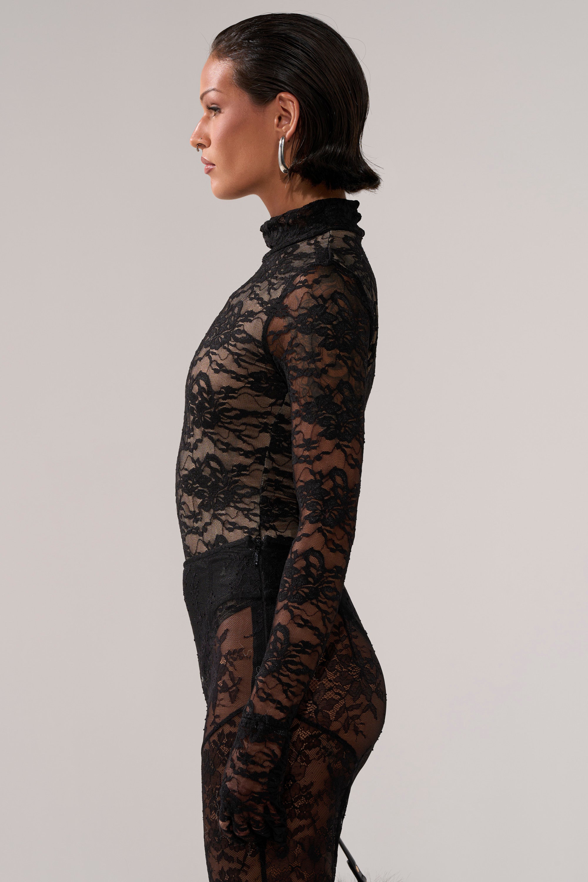A person with short dark hair and large hoop earrings wears the ALL THE ATTENTION LACE GLOVE BODYSUIT, featuring sheer floral lace, long sleeves, and a mock neckline, standing in profile against a plain light background.