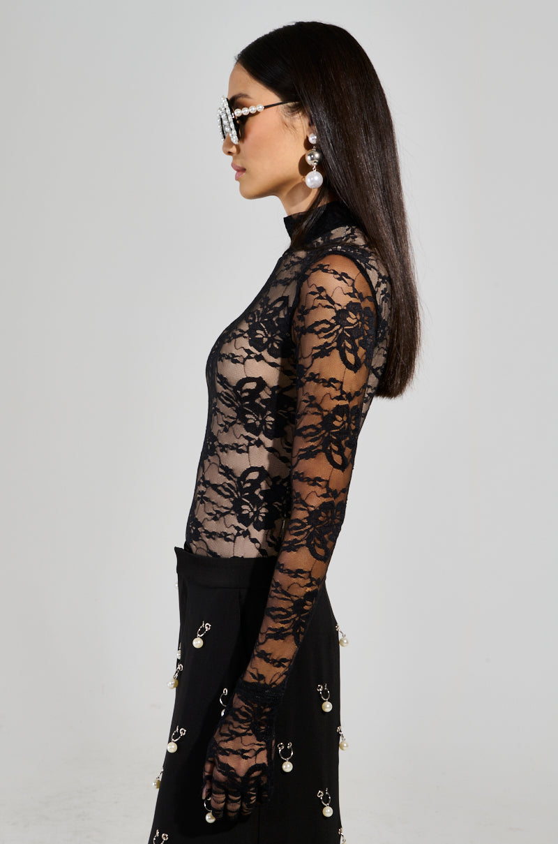 A woman with long dark hair, wearing oversized sunglasses and large pearl earrings, is dressed in the All The Attention Lace Glove Bodysuit, featuring a sheer floral lace design with long sleeves. She also sports a black skirt adorned with dangling pearl embellishments. The background is plain.