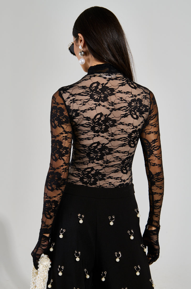 A person with long dark hair is wearing the "ALL THE ATTENTION LACE GLOVE BODYSUIT," characterized by its sheer floral lace design, paired with a black skirt adorned with pearls. They are holding a white, textured handbag and accessorized with large pearl earrings. The image captures them from behind.