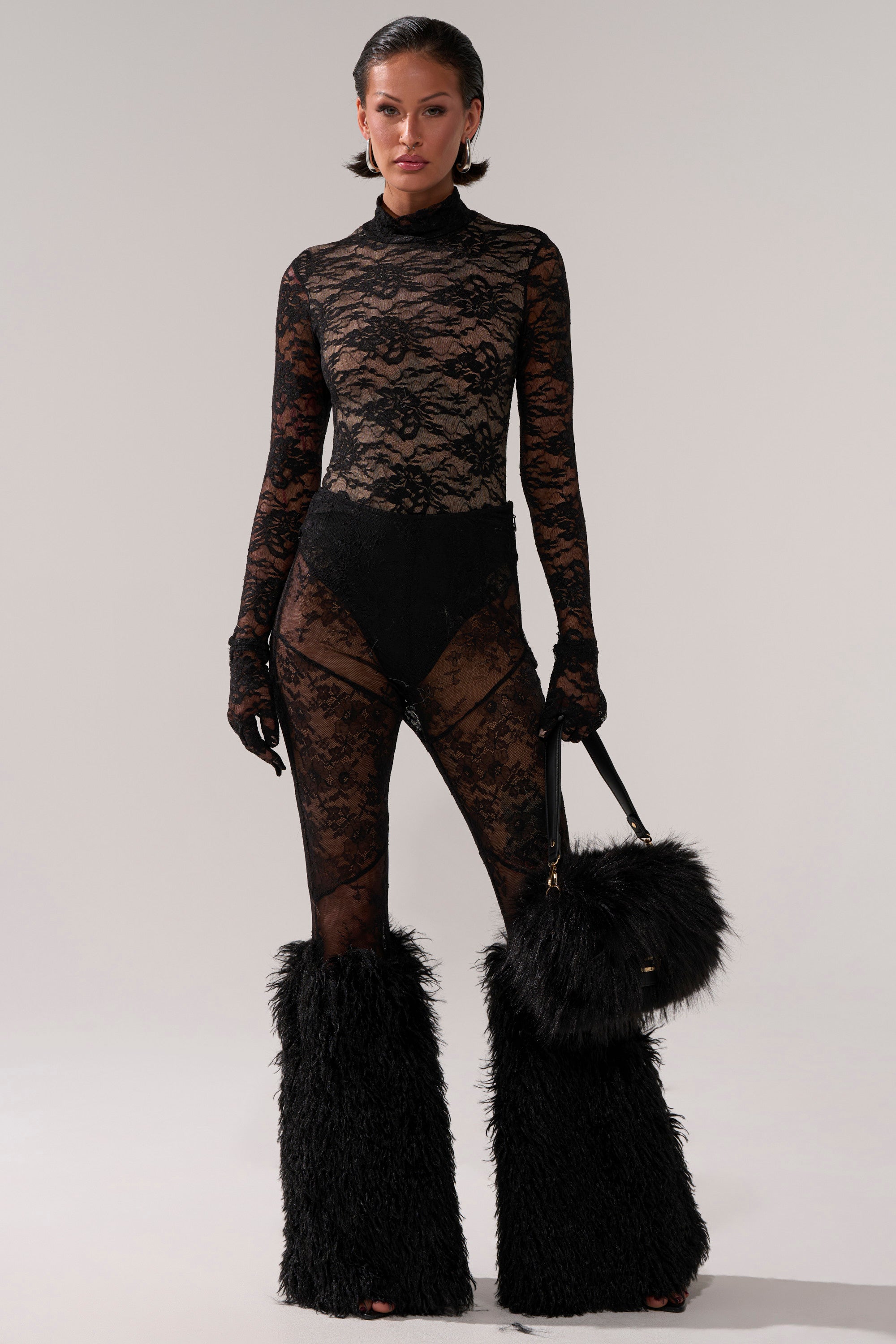 A woman poses against a plain background wearing the ALL THE ATTENTION LACE GLOVE BODYSUIT, paired with black underwear, long lace gloves, black furry leg warmers, and holding a large black furry handbag while looking at the camera.