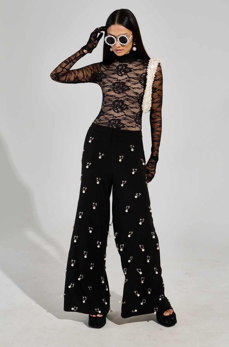 A person confidently poses in an ALL THE ATTENTION LACE GLOVE BODYSUIT, paired with embellished wide-leg pants. They complete the look with bold sunglasses and platform shoes, all set against a neutral studio backdrop.