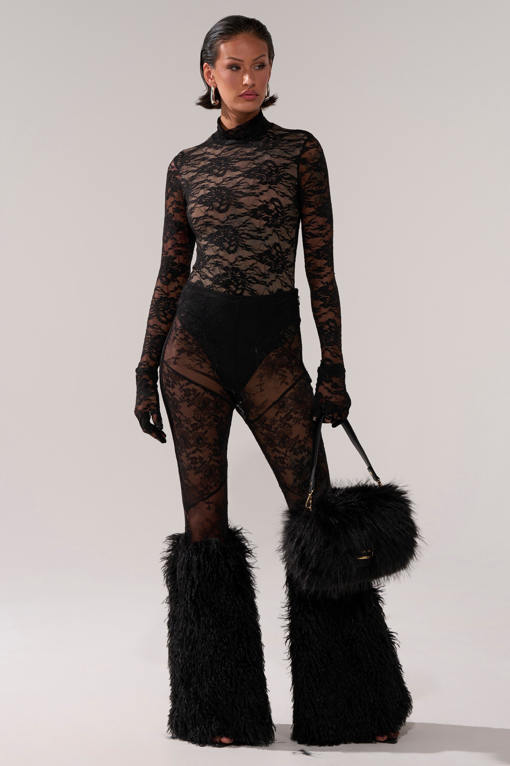 A woman models the ALL THE ATTENTION LACE GLOVE BODYSUIT with matching black underwear, a mock neckline, fluffy black leg warmers, statement earrings, and a black furry handbag against a plain light background.