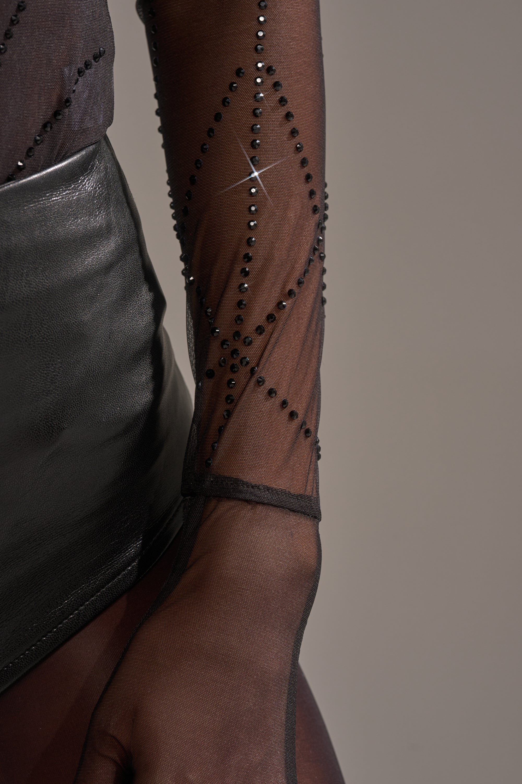 A close-up of a person wearing the SPIDER GIRL MESH BODYSUIT, a sheer black mesh outfit with rhinestone details, paired with a shiny black leather skirt against a plain, neutral background.