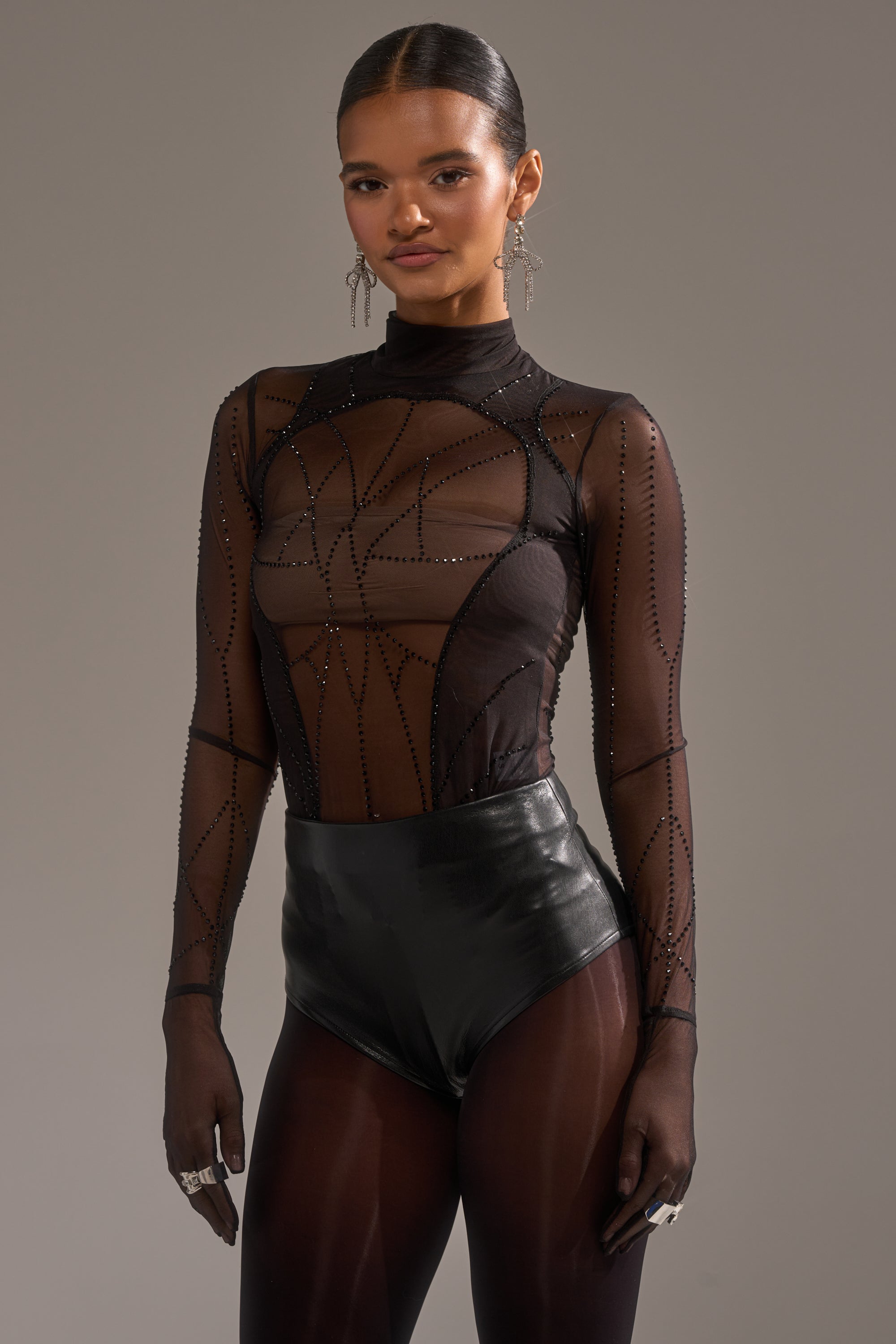 A woman poses confidently in the SPIDER GIRL MESH BODYSUIT, a sheer black mesh piece with geometric patterns, styled with black high-waisted bottoms, statement earrings, rings, and gloves against a neutral background.