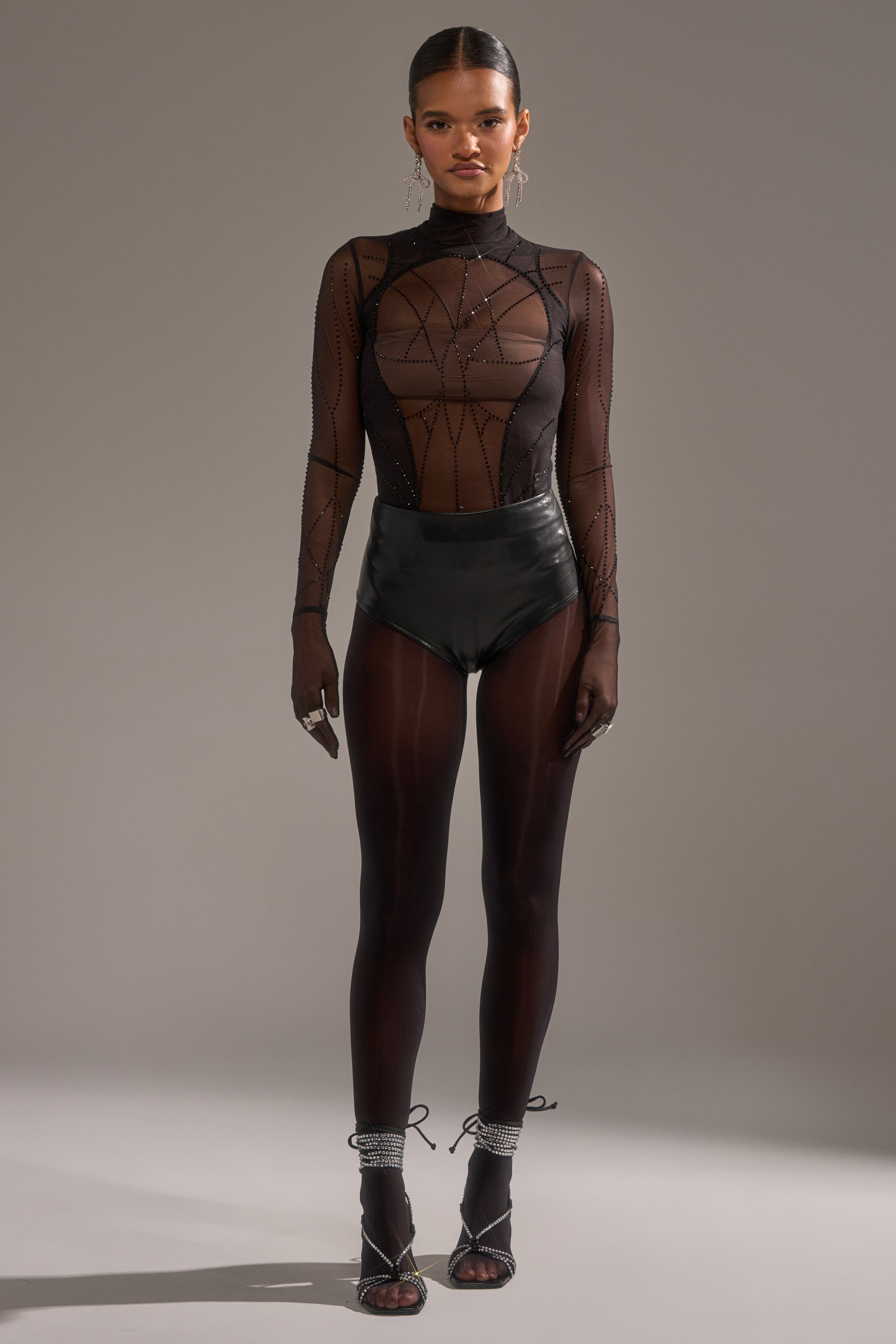 A woman poses against a plain background in the SPIDER GIRL MESH BODYSUIT featuring geometric patterns, paired with high-waisted black shorts, sheer tights, strappy heels, and statement earrings.