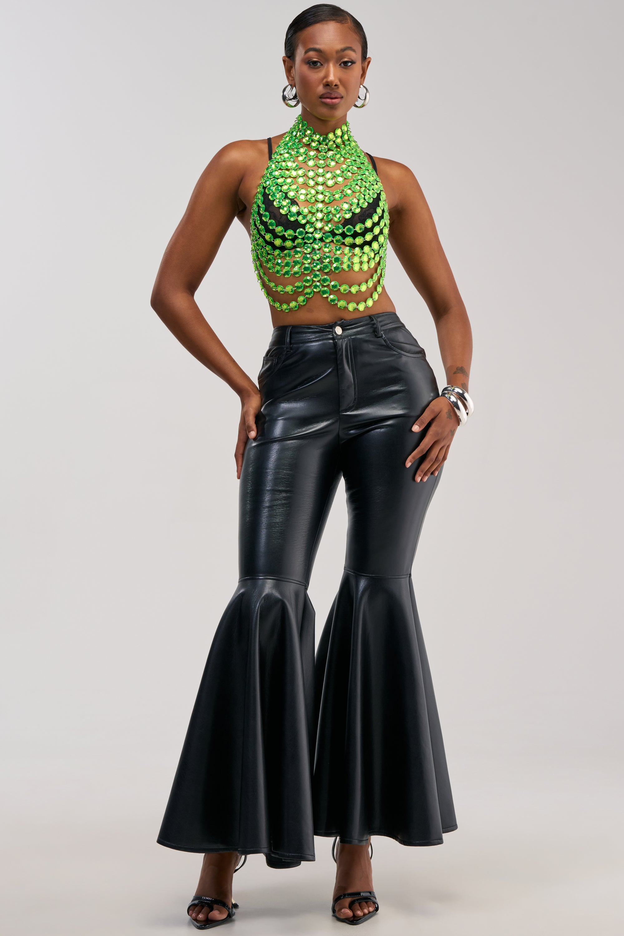 A person poses confidently in a green sequin halter top and the FLY AWAY WITH ME FAUX LEATHER FLARE TROUSER, featuring a flattering high-rise fit, paired with heels and hoop earrings against a plain background—perfect night out styling.