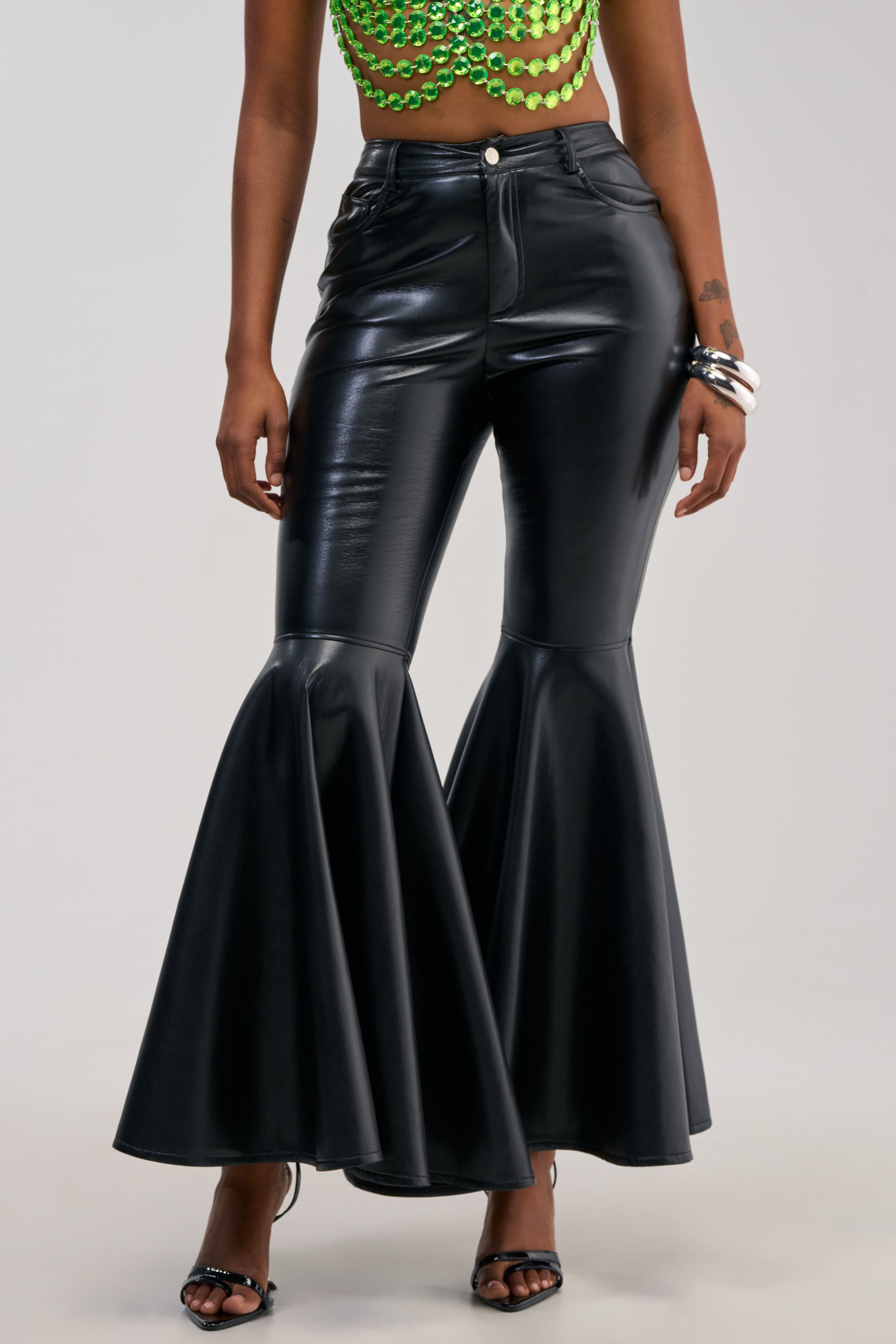 A person stands against a plain background, styled for the ultimate night out in the FLY AWAY WITH ME FAUX LEATHER FLARE TROUSER—a pair of high-waisted, shiny black faux leather flares with wide legs—paired effortlessly with a green beaded top, black sandals, and silver jewelry.