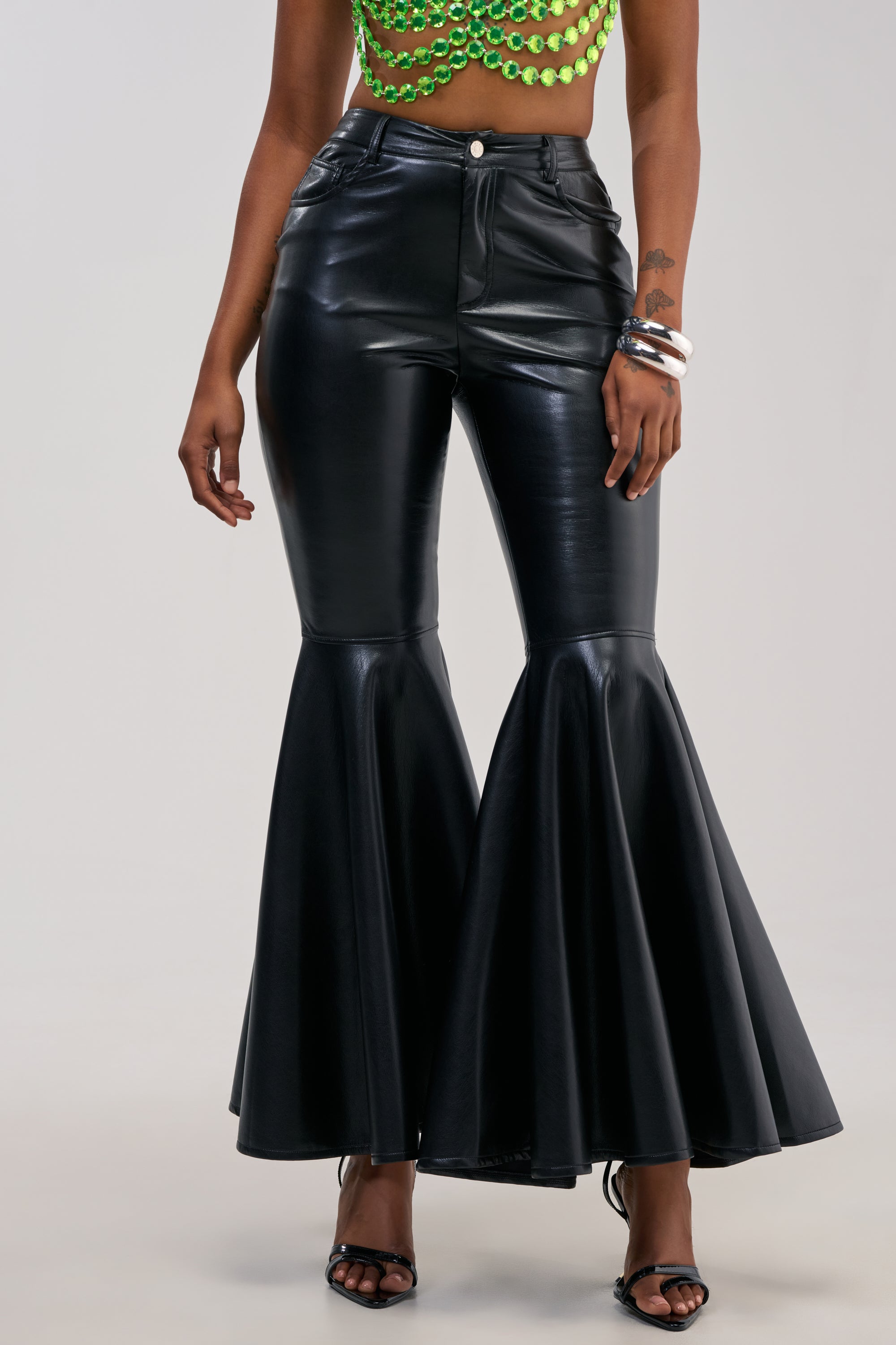 Wearing the FLY AWAY WITH ME FAUX LEATHER FLARE TROUSER in black with a high-rise fit, paired with a green beaded crop top, black heeled sandals, and silver bracelets, this look stands out against a plain background—ideal for chic night-out styling.