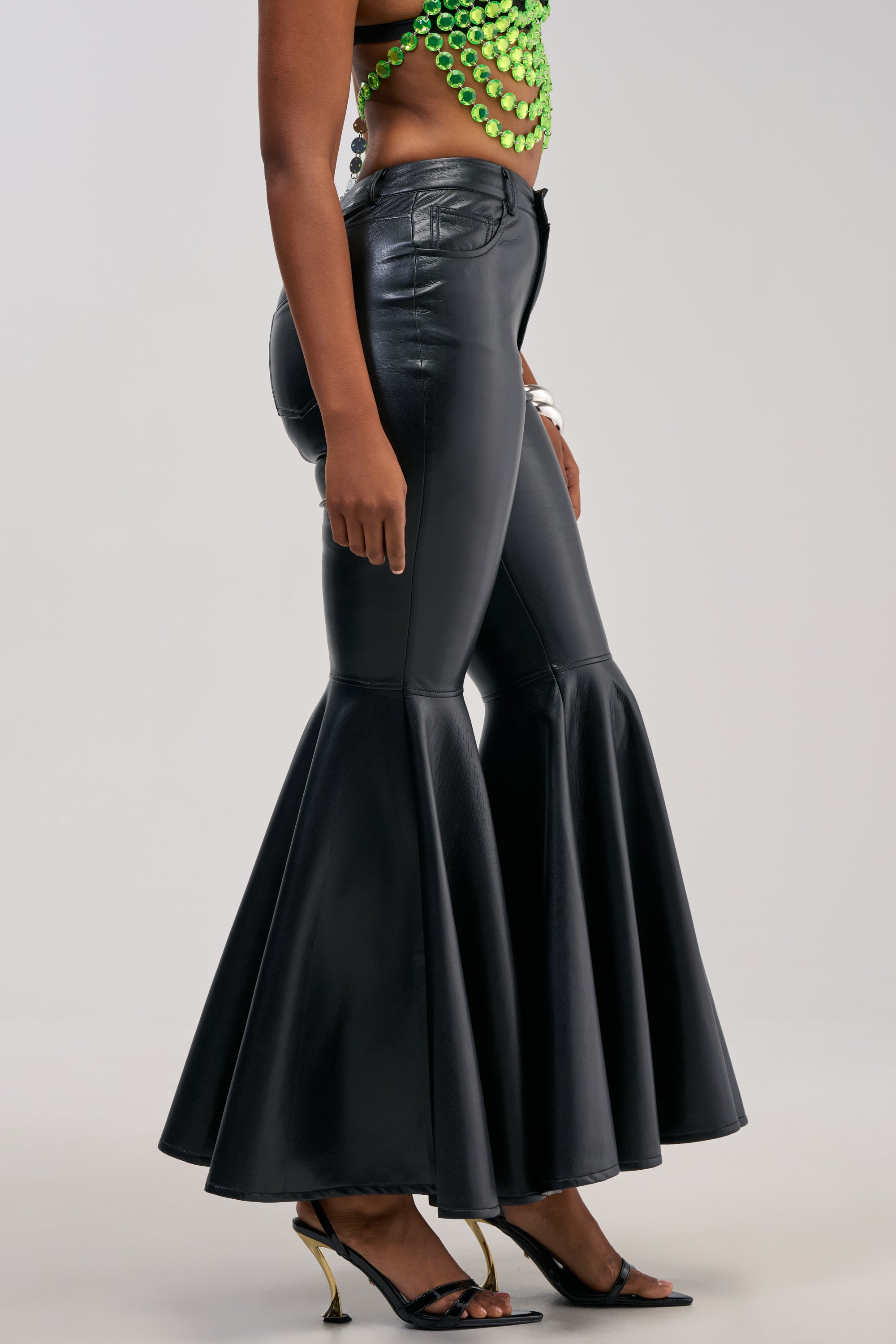 Wearing the FLY AWAY WITH ME FAUX LEATHER FLARE TROUSER in black, styled with a green beaded crop top and black open-toe heels featuring gold heels, this look is perfect for a night out. The image captures the person from the thighs down against a plain background.