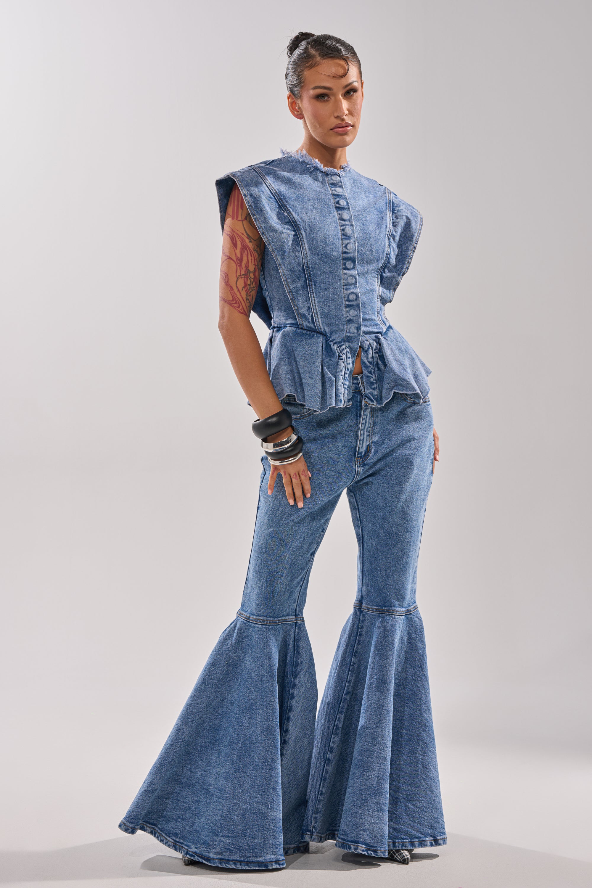 A person stands confidently in the DONT PLAY WITH MY HEART BELL BOTTOM DENIM—high-rise, flared jeans—paired with a sleeveless, light-wash denim top with ruffled shoulders. Their hair is tied back and a tattoo shows on their upper arm.