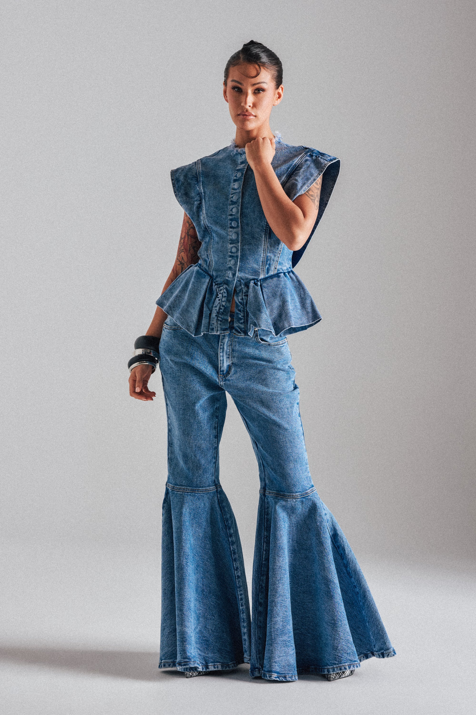 A person stands confidently in DONT PLAY WITH MY HEART BELL BOTTOM DENIM, featuring flared jeans and a sleeveless, structured top. Tattoos cover one arm and chunky bracelets complete the look against a plain light gray background.