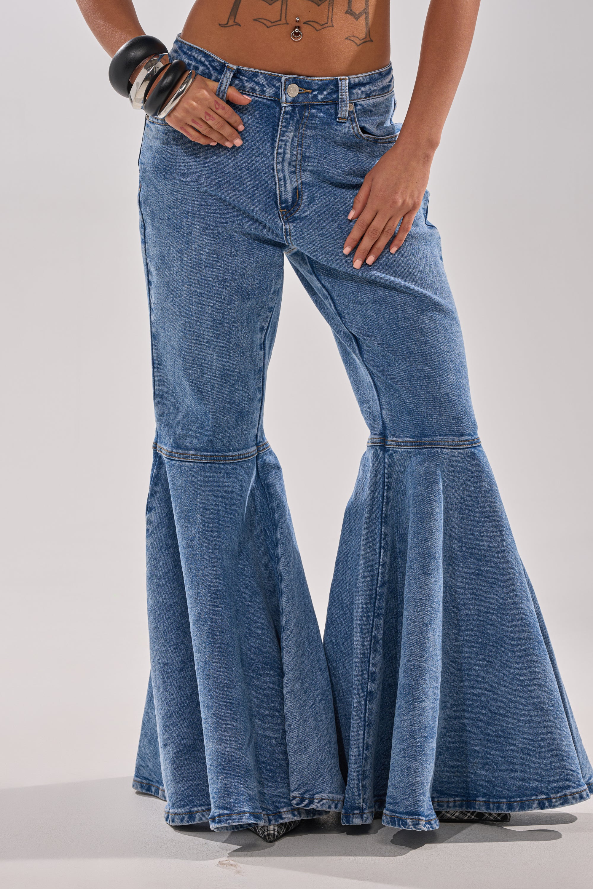 Someone models the DONT PLAY WITH MY HEART BELL BOTTOM DENIM—high-rise, blue jeans with dramatic wide flares—styled with chunky black bracelets and a midriff showcasing tattooed designs.