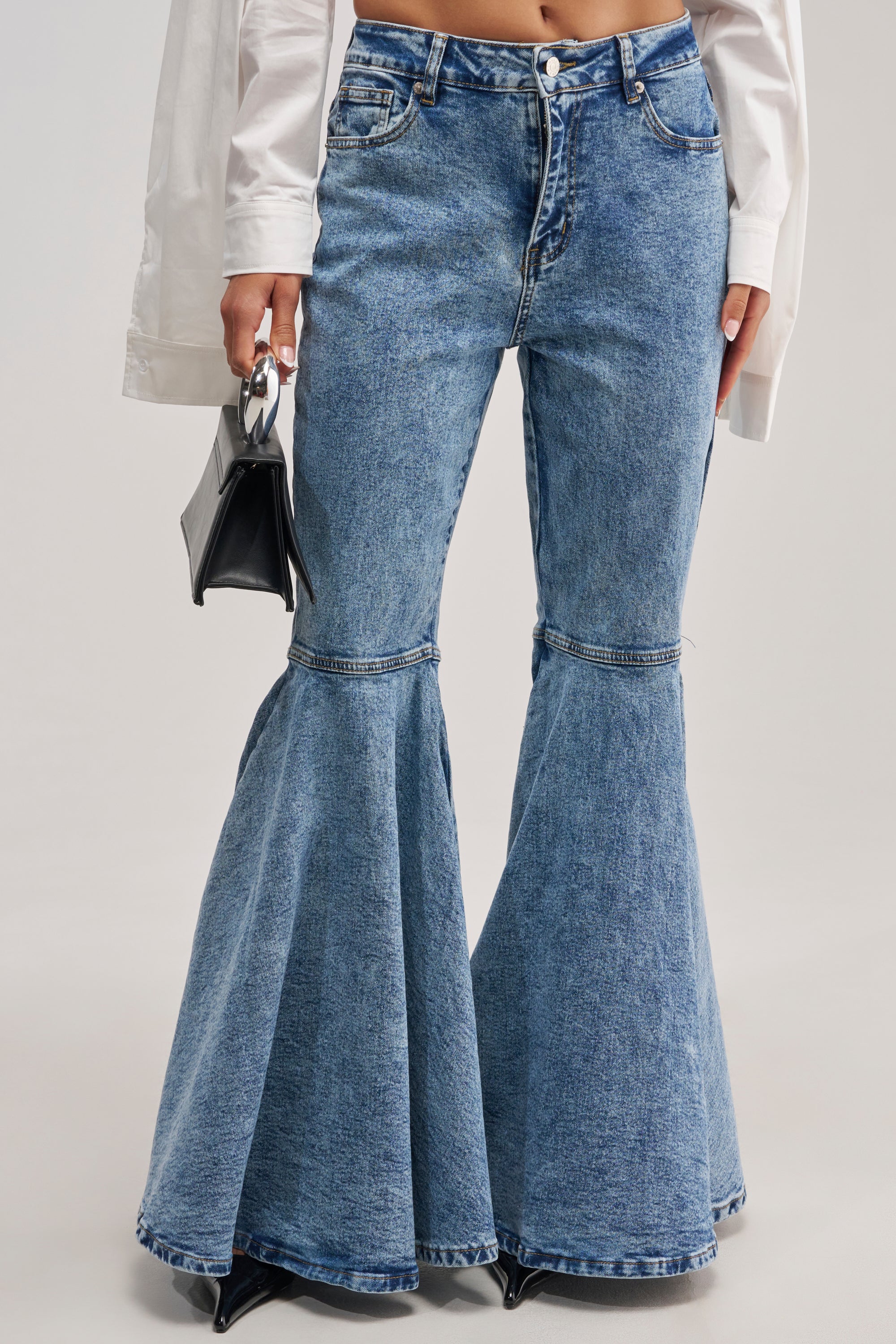Wearing the DONT PLAY WITH MY HEART BELL BOTTOM DENIM—a pair of high-waisted blue jeans with dramatic wide bell bottoms and visible seam details—styled with a white long-sleeve shirt and a small black handbag for a bold streetwear look.