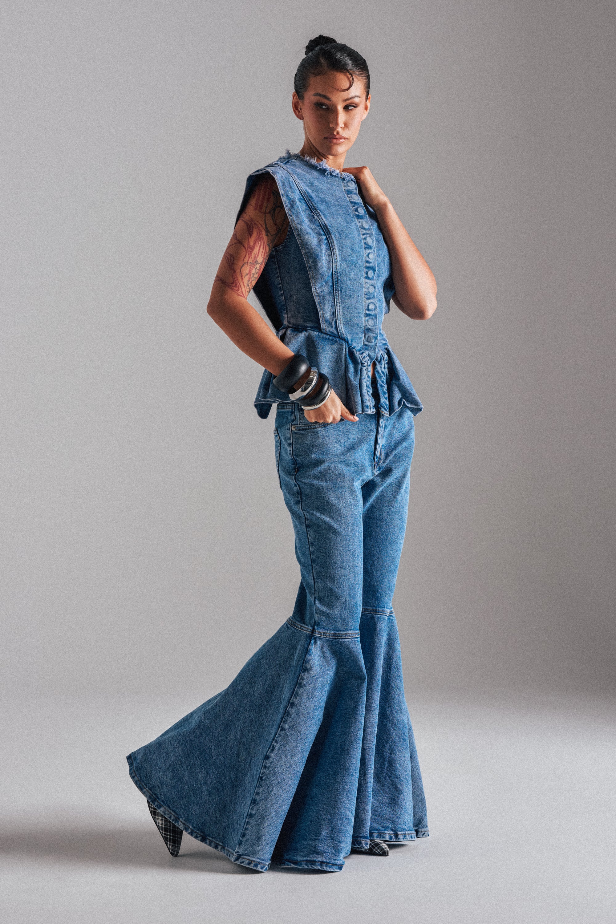 A woman models the DONT PLAY WITH MY HEART BELL BOTTOM DENIM jeans, paired with a sleeveless denim top. She showcases arm tattoos and bracelets while posing against a plain, light-colored background.