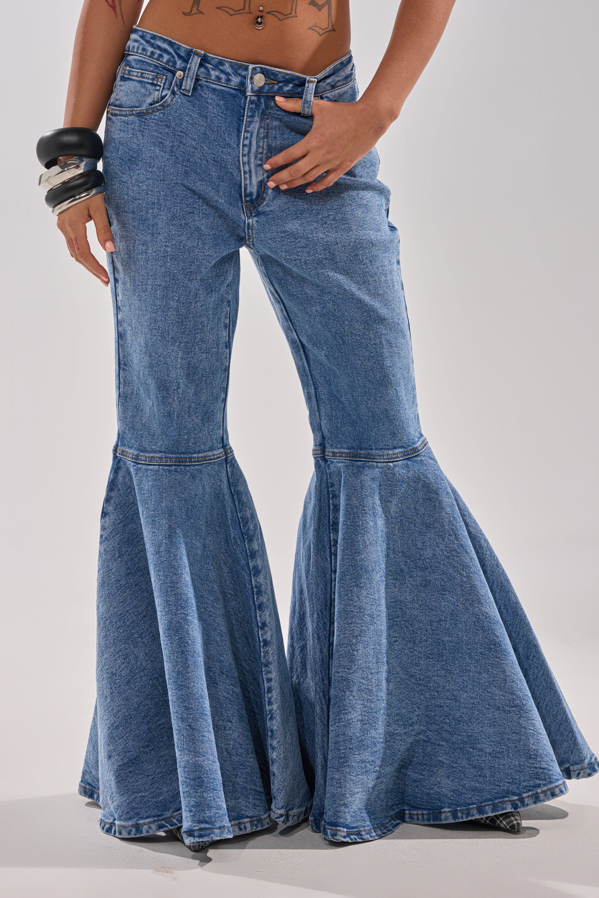 Wearing the DONT PLAY WITH MY HEART BELL BOTTOM DENIM, featuring high-waisted blue flared jeans with exaggerated wide bell-bottoms, paired with chunky bracelets and a tattooed midriff.