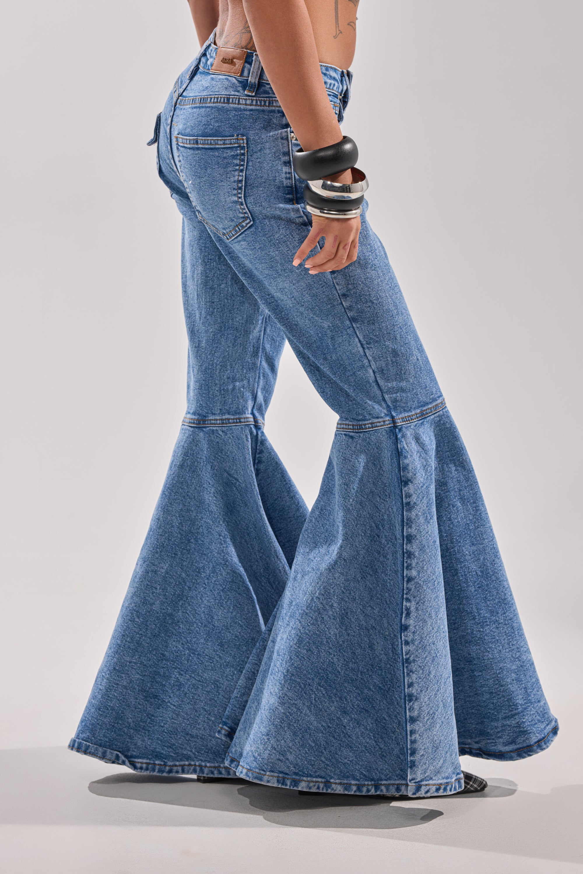 Wearing DONT PLAY WITH MY HEART BELL BOTTOM DENIM, a person stands with one leg slightly forward, showcasing the dramatic flare of these high-rise blue jeans and accessorized with black bracelets.