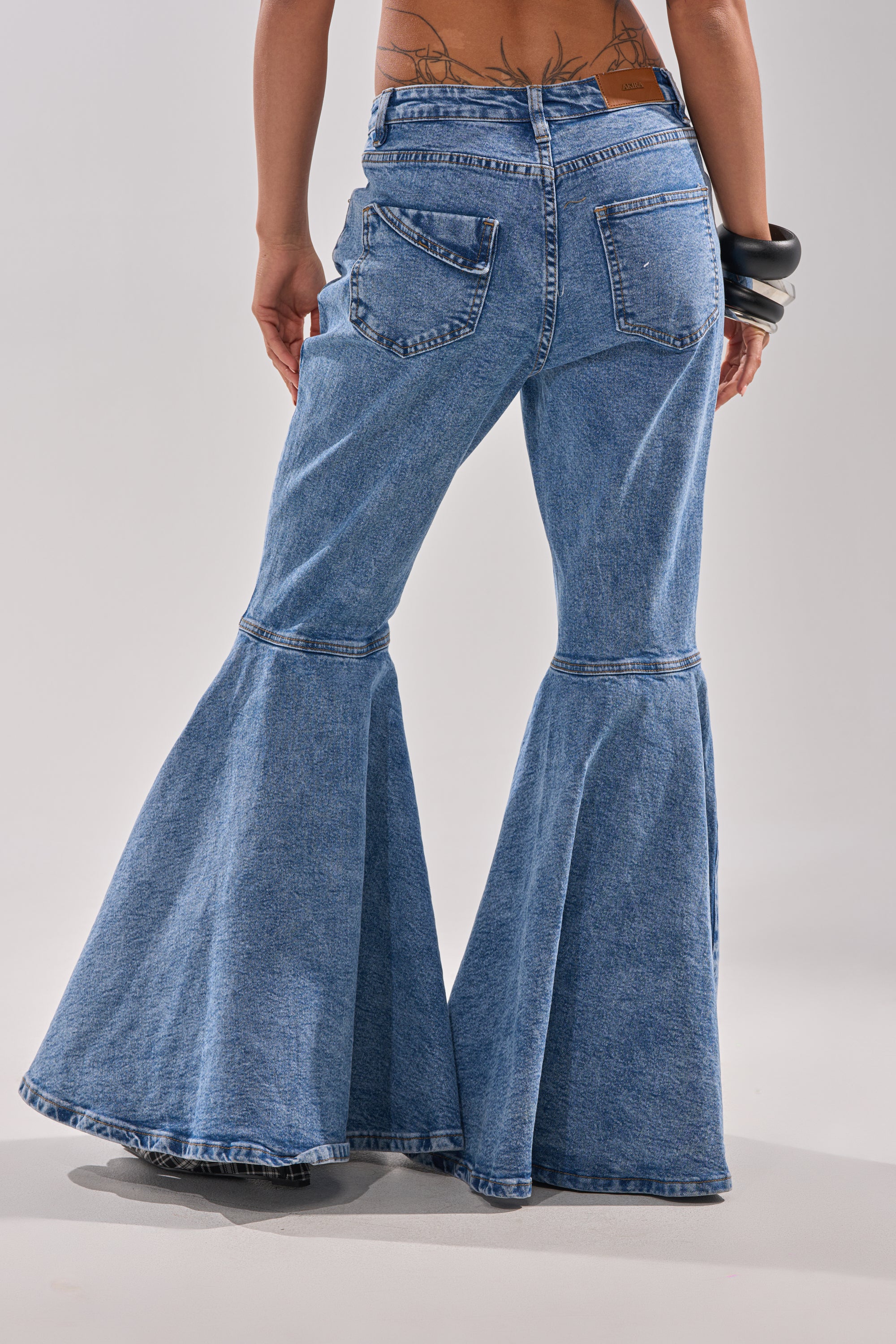 A person is seen from the back wearing DONT PLAY WITH MY HEART BELL BOTTOM DENIM—blue, high-rise jeans with two back pockets and wide bell bottoms. Tattoos decorate their lower back, and they accessorize with large black bangles.