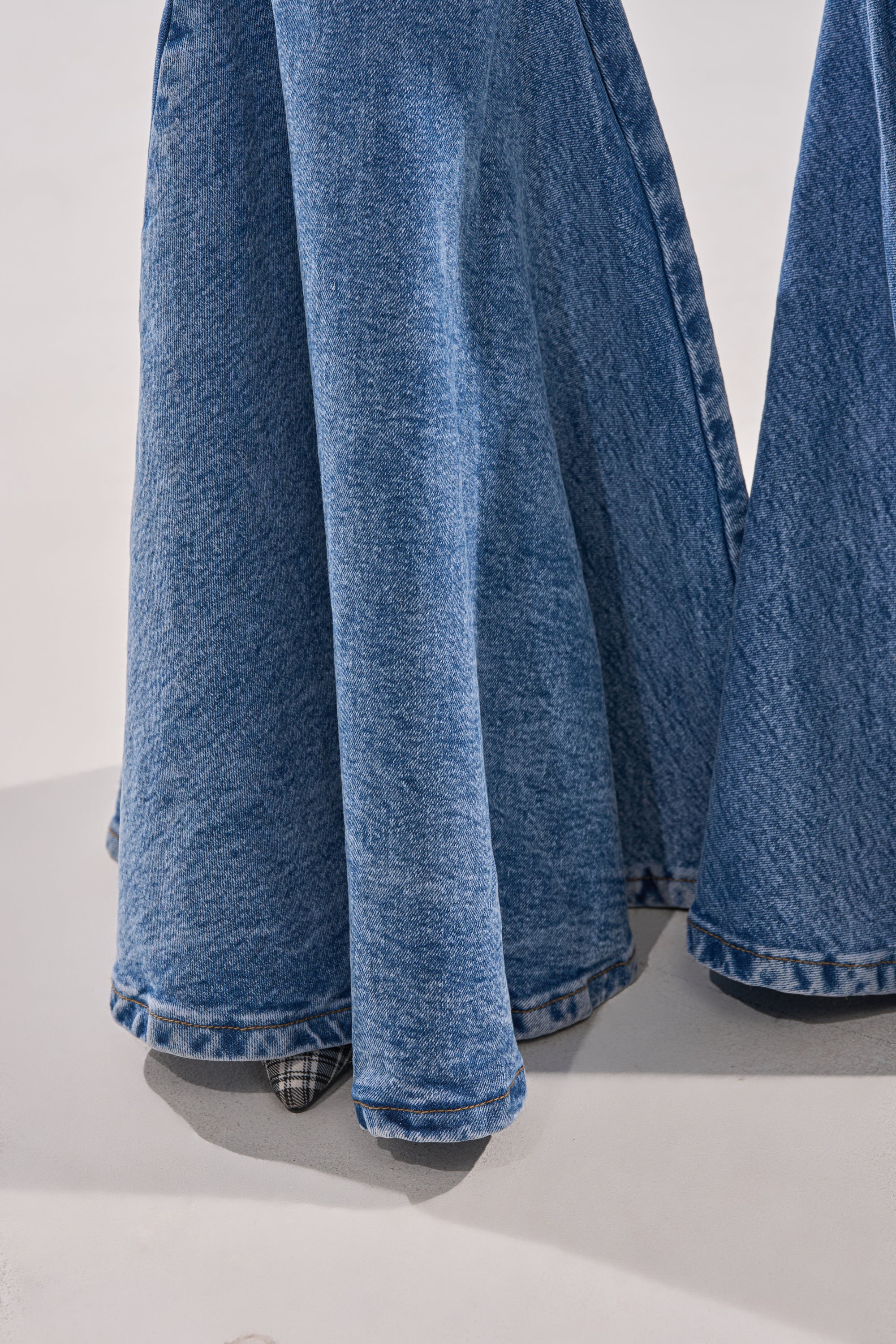 A close-up of two DONT PLAY WITH MY HEART BELL BOTTOM DENIM jeans just above the ground, with a plaid shoe peeking out from beneath the left bell bottom, against a smooth, light background.