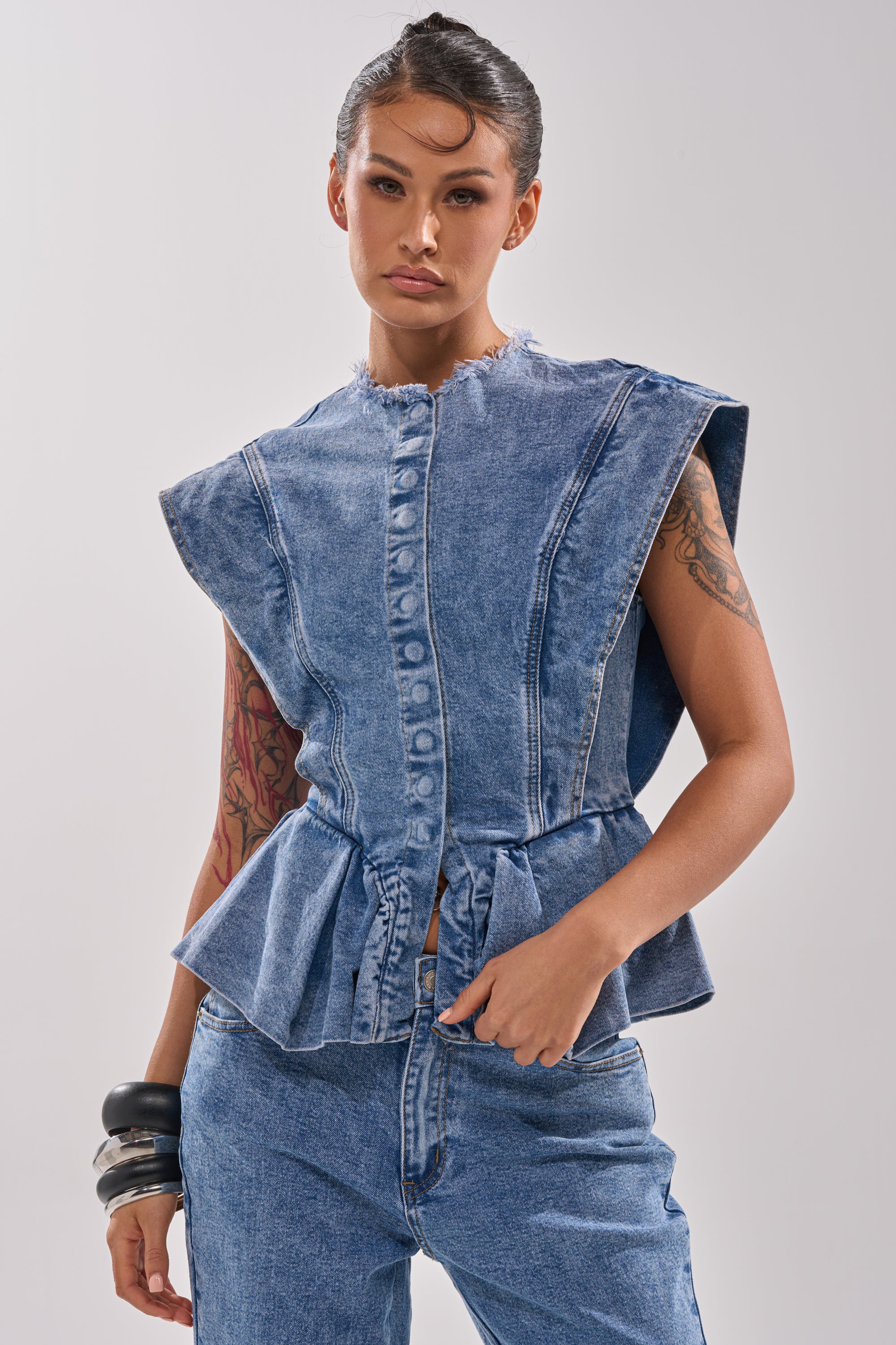 A woman with tattoos and chunky bracelets poses in the DONT PLAY WITH MY HEART DENIM BLOUSE, featuring a sleeveless design and ruffled peplum hem, paired with matching jeans against a plain light background. Her hair is styled back.